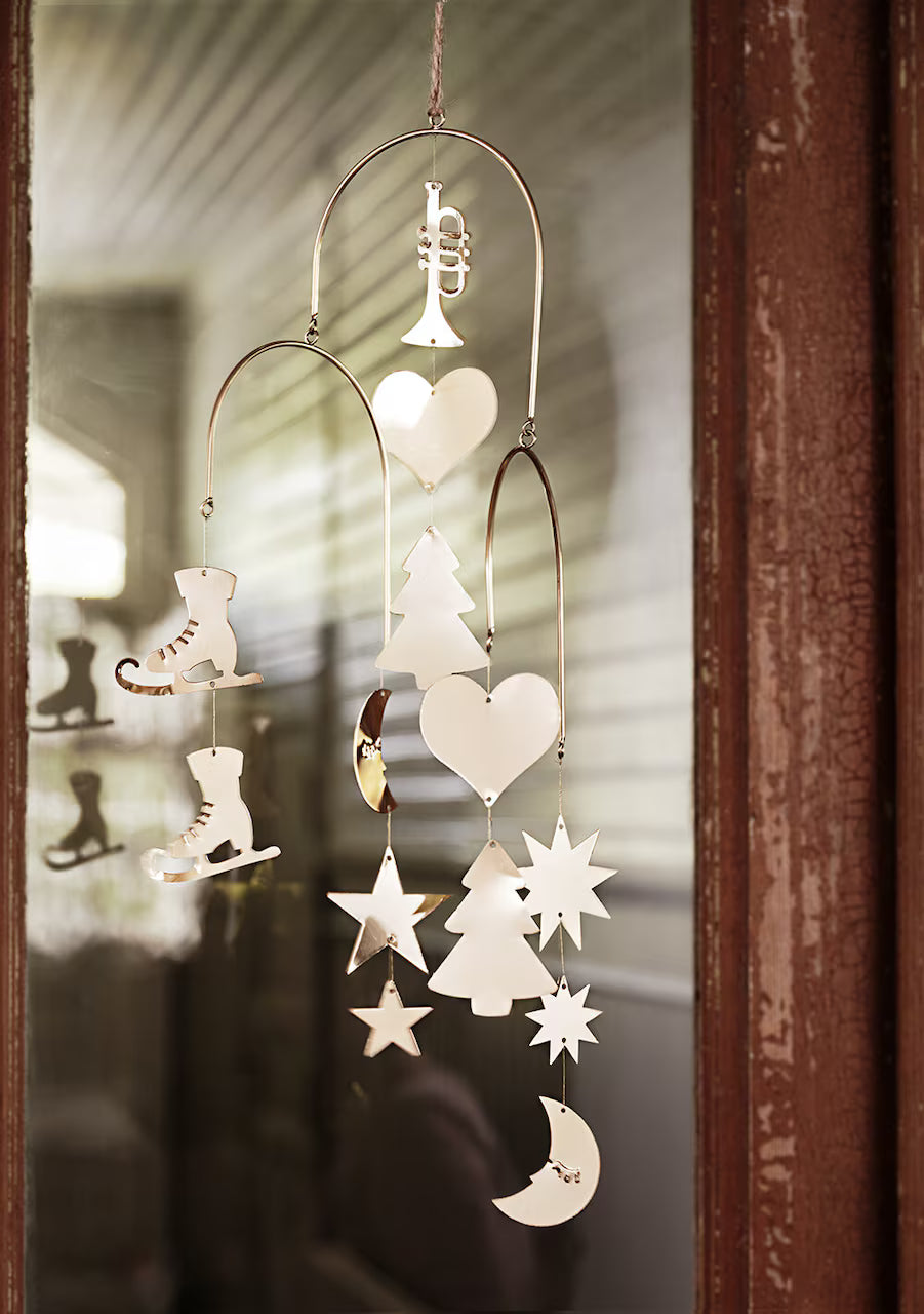 A Mobile Ornament in brass from Strömshaga hangs on a glass door, featuring cutouts of a trumpet, ice skates, trees, hearts, stars, and a crescent moon—classic Christmas motifs reminiscent of timeless ornaments. The reflection on the glass reveals similar shapes inside the house.