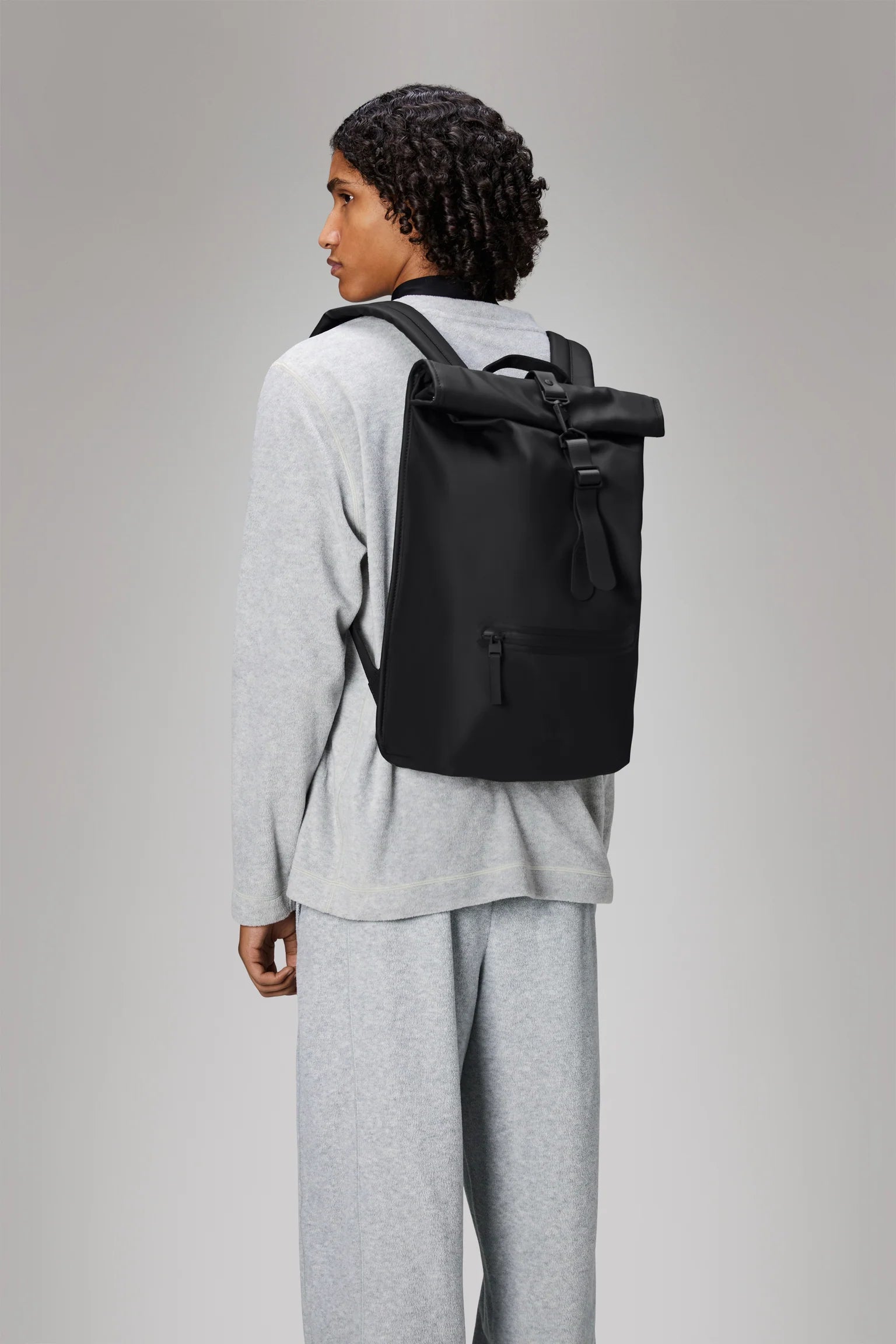 A person dressed in a light gray sweatshirt and pants, facing slightly away, carries the Rains Rolltop Rucksack Backpack in Black by Rains against a plain, light background.