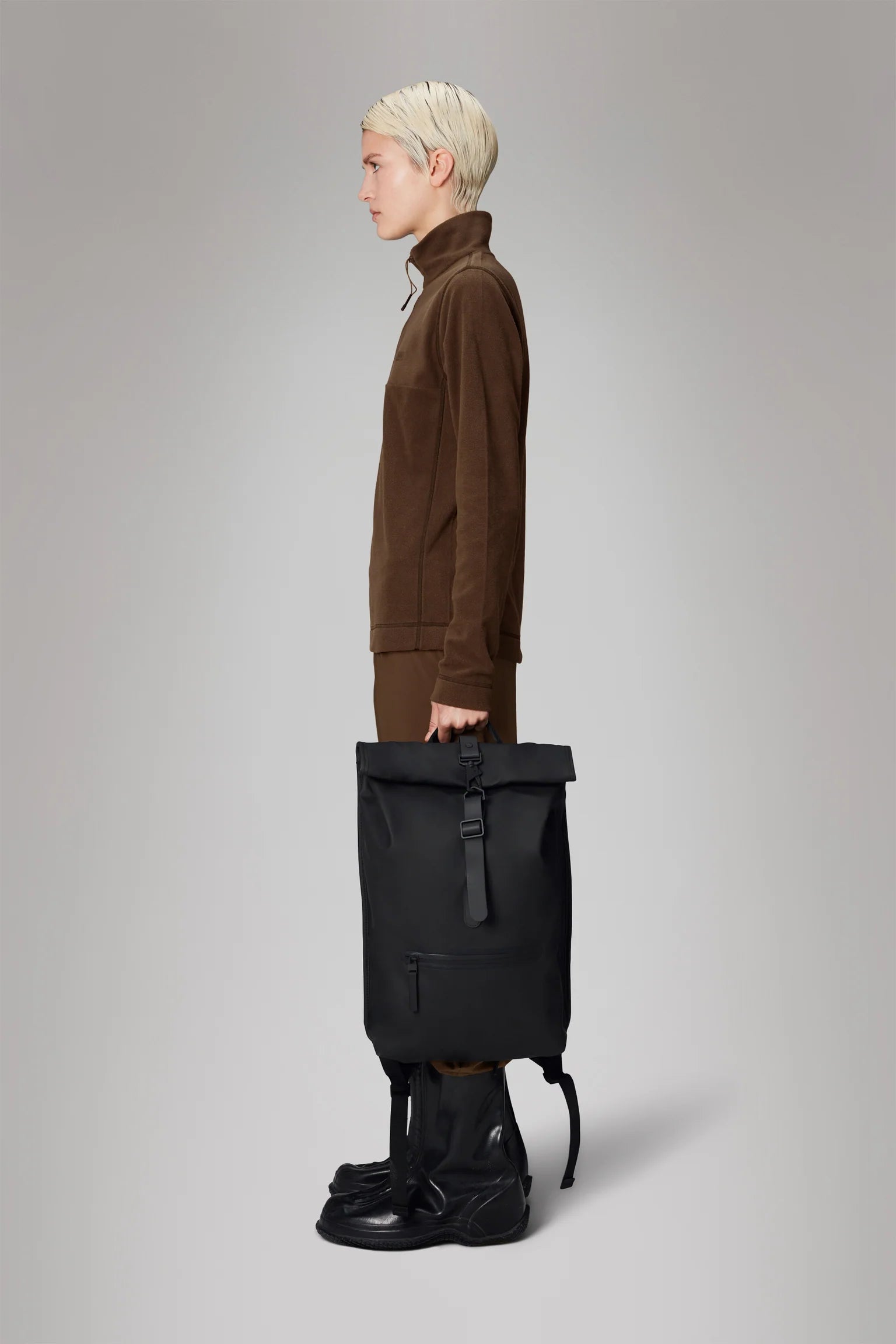 A person with short blond hair wearing a brown outfit and tall black boots stands in profile, holding the Rains Rolltop Rucksack Backpack in Black by Rains against a plain gray background.