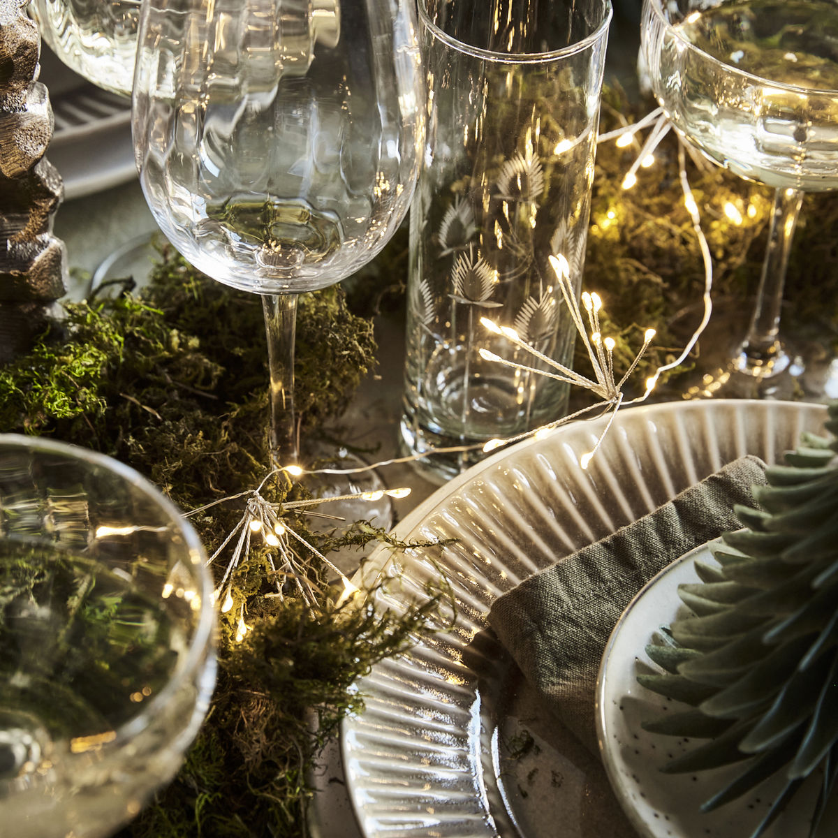 A festive table setting features glassware, plates, green napkins, moss, a green tree-shaped candle, and House Doctor String Lights, Glow LED, Silver Finish 1.5m woven through for a sparkling touch.