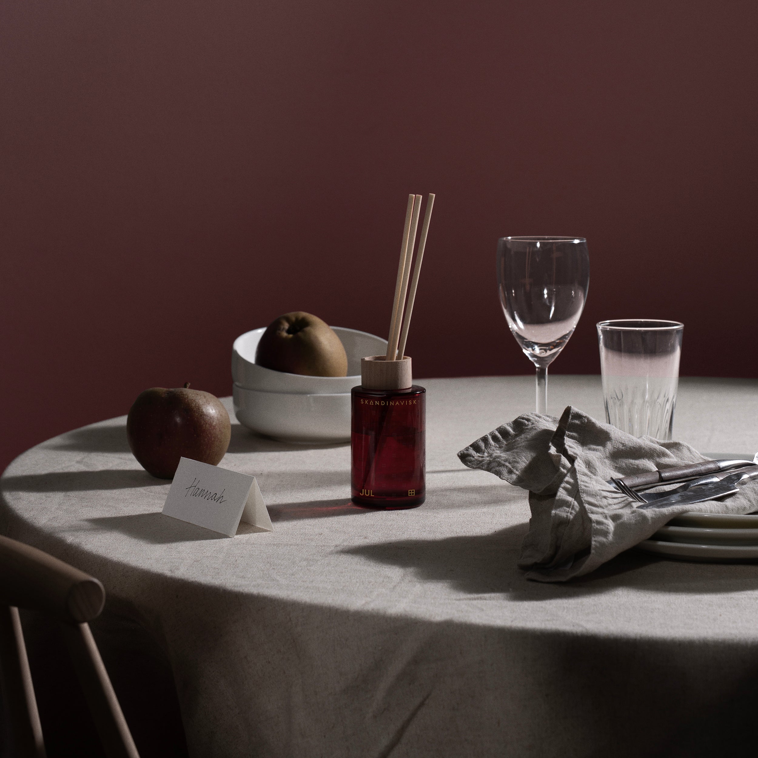 A round table with a beige tablecloth displays apples, a place card, the Skandinavisk Jul Scent Diffuser (100 ml), stacked plates, glassware, and a folded napkin against a dark pink background.