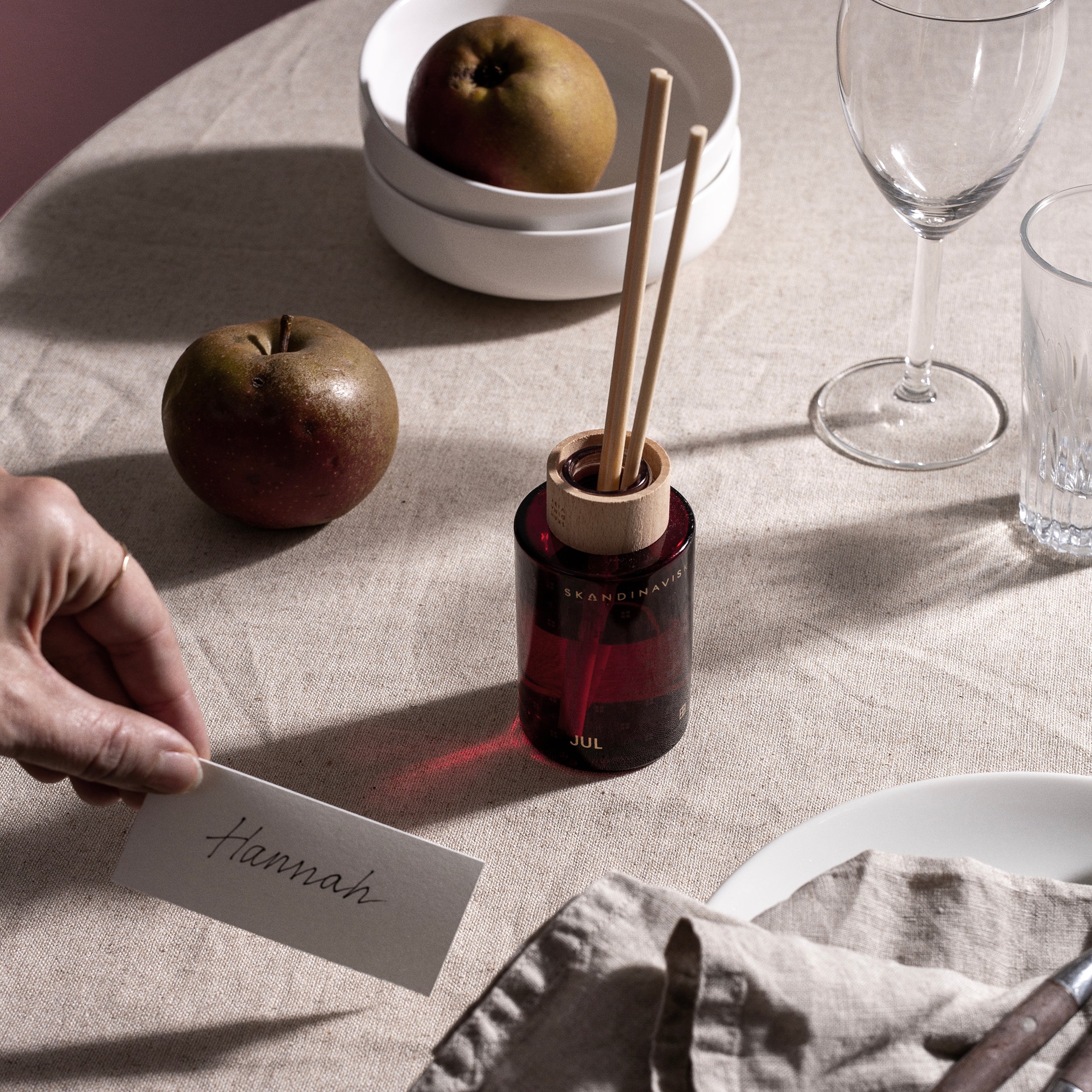 A hand places a card labeled Hannah on a linen table with apples, glassware, a napkin, and the Skandinavisk Jul Scent Diffuser (100 ml), capturing the cozy warmth of a Scandinavian Christmas.