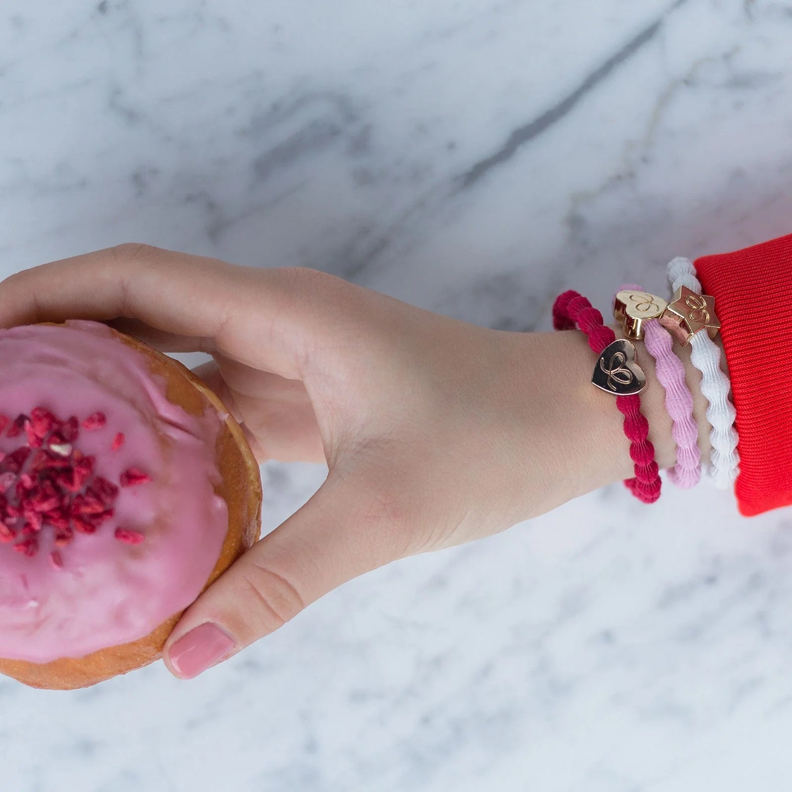 A hand with pink-painted nails holds a pink-iced donut, wearing the ByEloise Bangle Band Gold Star · Crisp White and colorful charm bracelets, all set against a marble background.