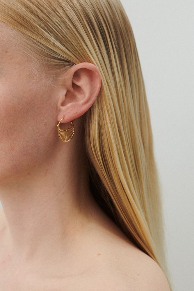 A close-up of a person with long blonde hair wearing a stunning gold Daylight Earring by Pernille Corydon in their left ear.