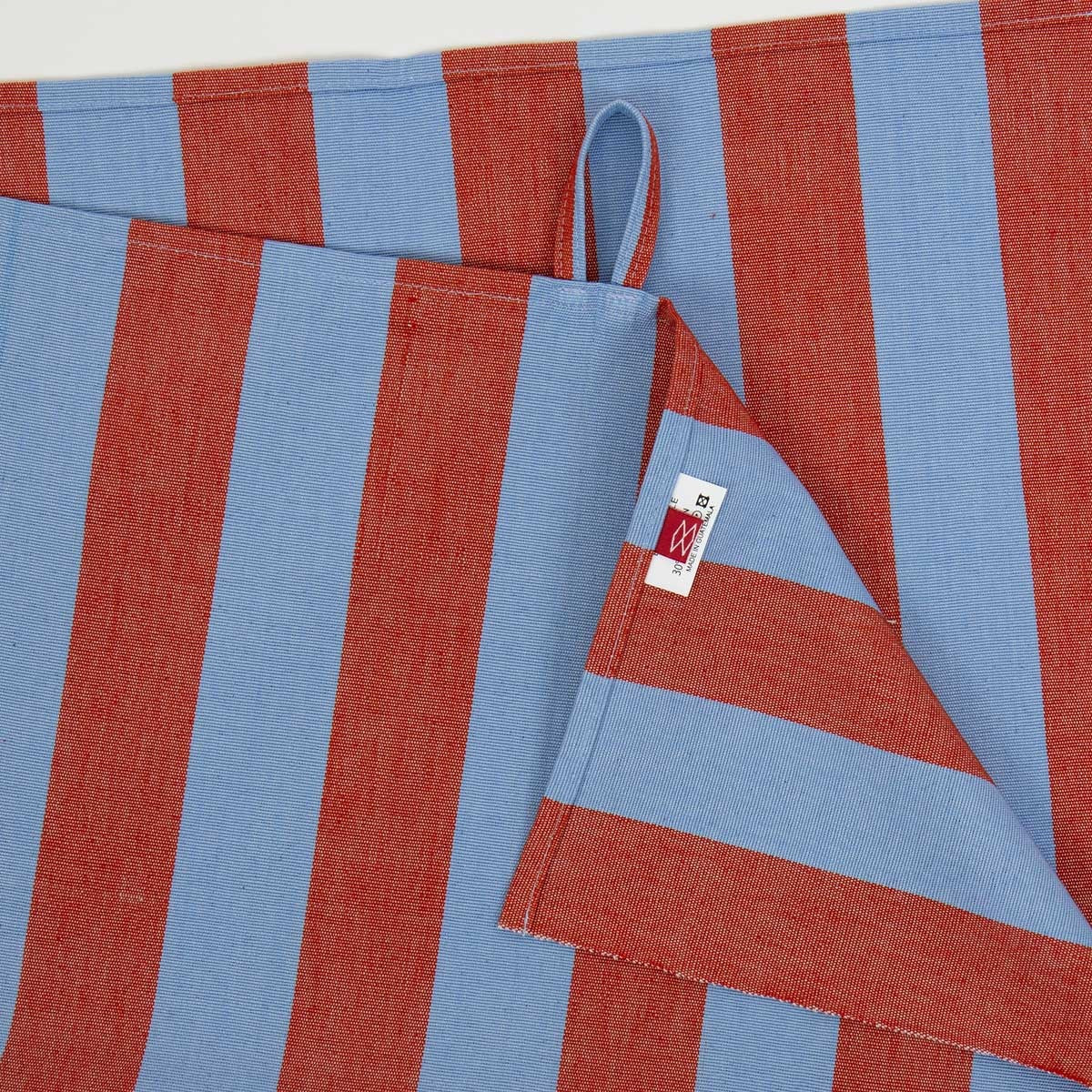 Two “Nora Striped in Blue & Red” organic cotton tea towels from A World of Craft are layered against a white background. One towel is partially folded to showcase its corner loop and a white tag with red and blue writing. Handwoven and fair trade, these Swedish-inspired towels add a stylish touch to any setting.