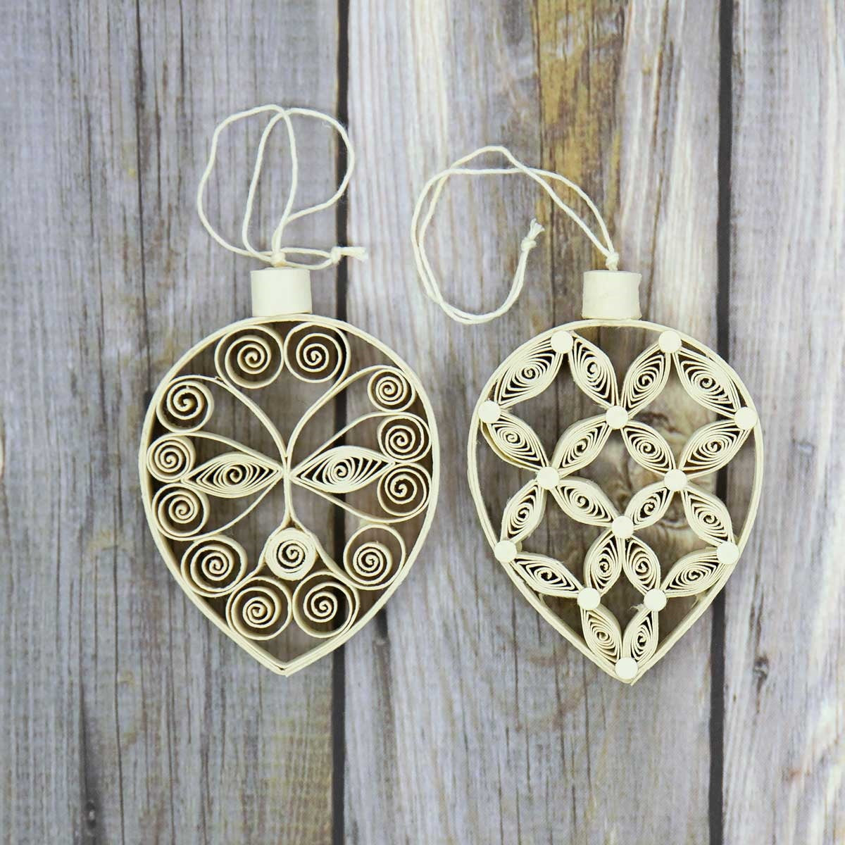 Two handcrafted Quilled Leaf Tree Christmas Ornaments by A World of Craft, each made from coiled and shaped beige paper strips that form intricate quilled patterns, hang against a wooden background. This 2-pack offers beautiful handmade decorations for any space.