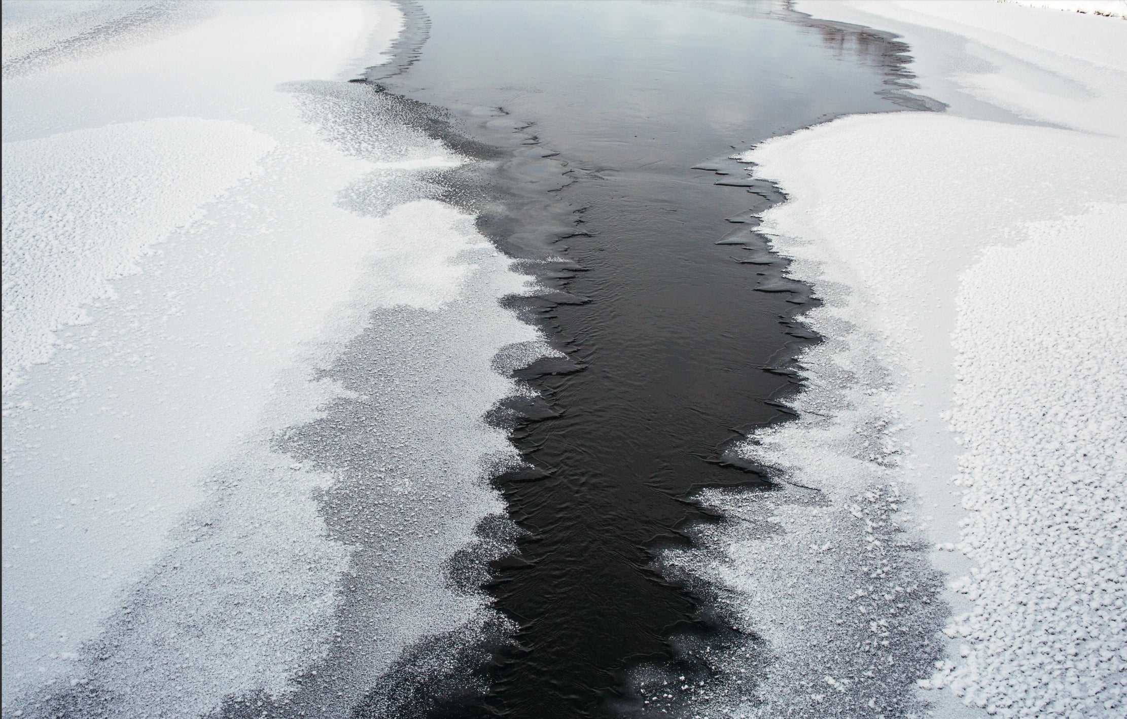 A river, partially iced and snow-covered, winds through a Nordic winter scene—captured in "Genuine – Handmade Beauty from the North" by Cozy Publishing. The dark water contrasts with frosty banks and jagged ice along the shore.