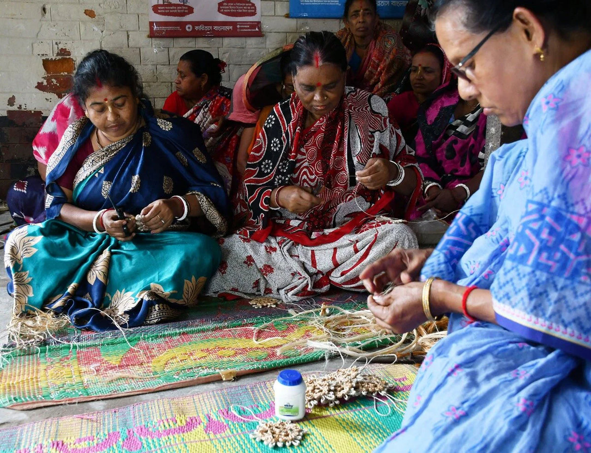 Women sit on colorful mats weaving and crafting with natural fibers, focused on their work. They wear bright, patterned sarees and are surrounded by materials in a communal indoor setting. Among their creations are rustic Christmas décor pieces—such as the Strömshaga Nisse Santa Ornament Handmade in Jute, featuring red and off-white details—blending tradition with festive artistry.