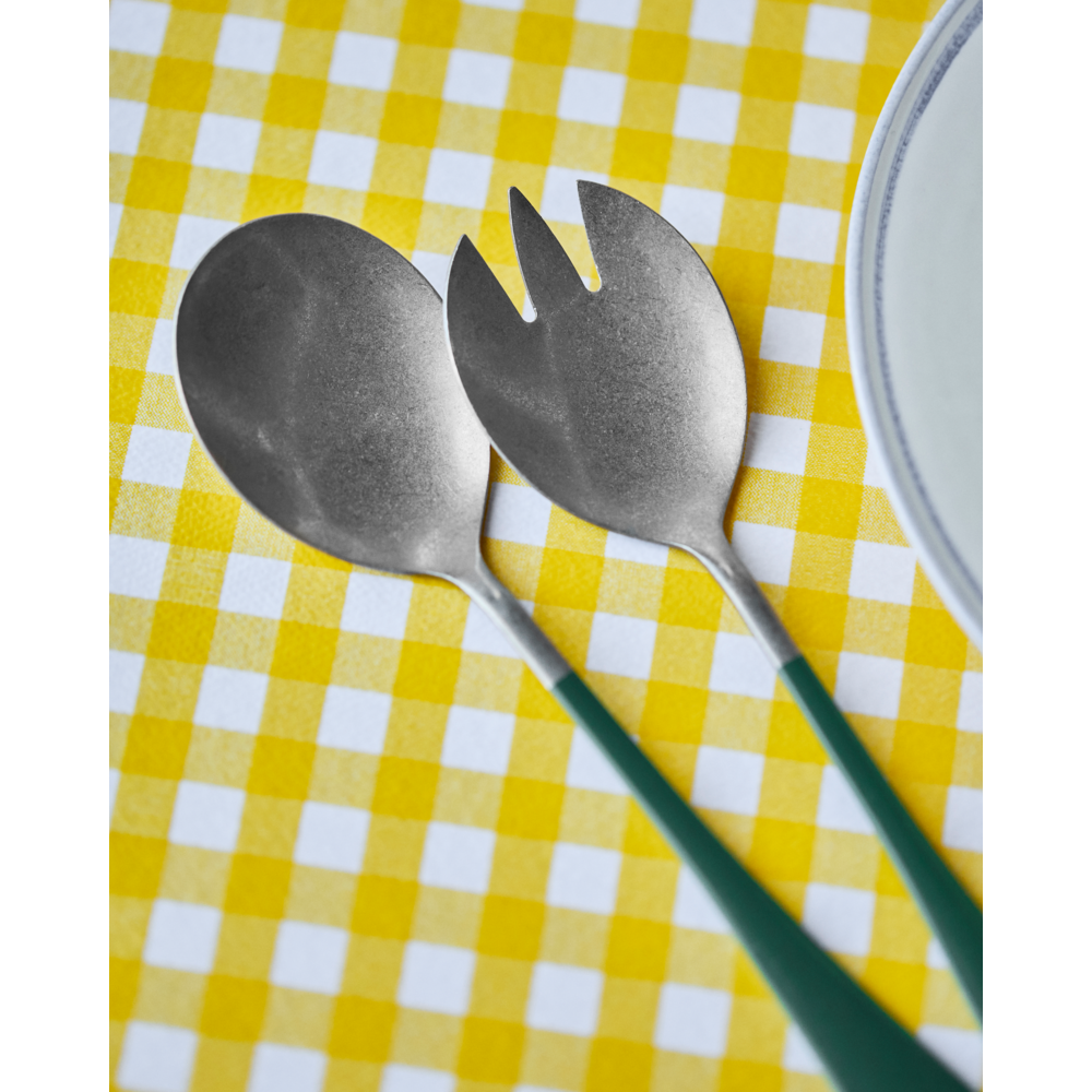 A pair of Nicolas Vahé Salad Servers in Stainless Steel, each with a green handle and antique silver finish—one spoon and one spork—rest on a yellow and white checkered tablecloth beside a white plate with a blue rim.