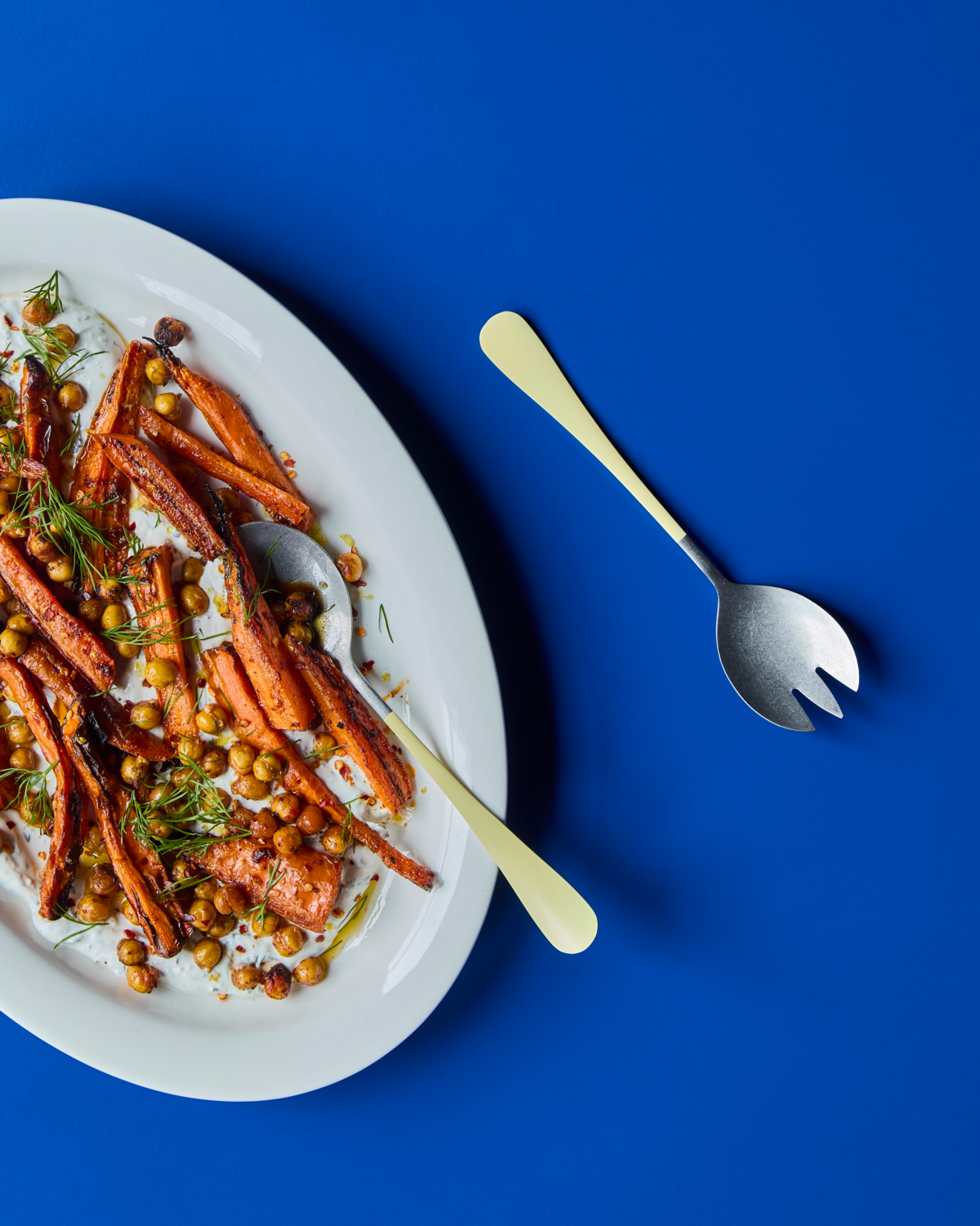 A white oval plate with roasted carrots, chickpeas, herbs, and creamy sauce sits on a blue surface. To the right are the Nicolas Vahé Salad Set in Stainless Steel, Yellow—fork and spoon arranged diagonally.