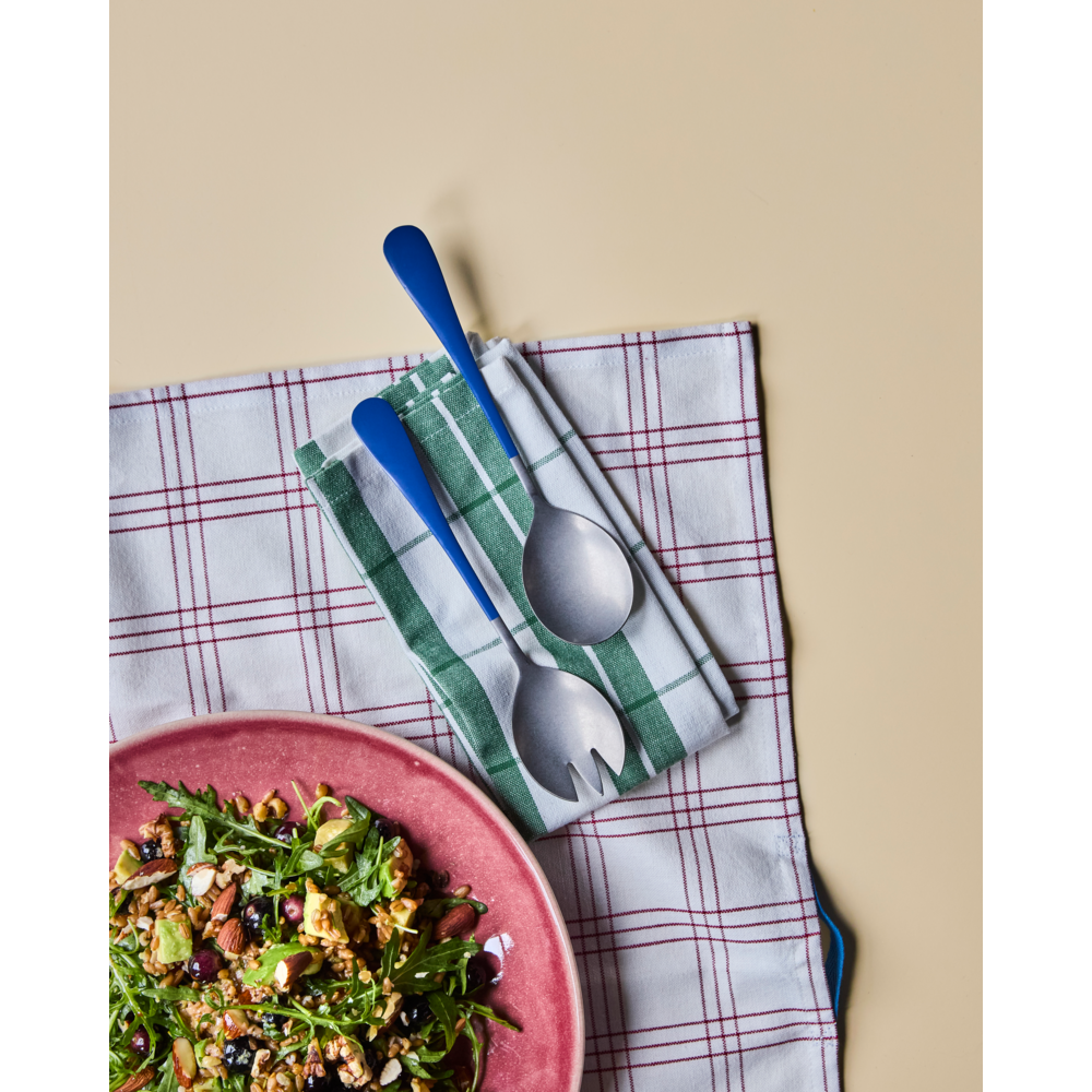 A pink plate with salad sits on a red-and-white checked cloth. Next to it are green-striped and checked napkins topped with the Nicolas Vahé Salad Set in Stainless Steel, Blue, all arranged on a beige surface.