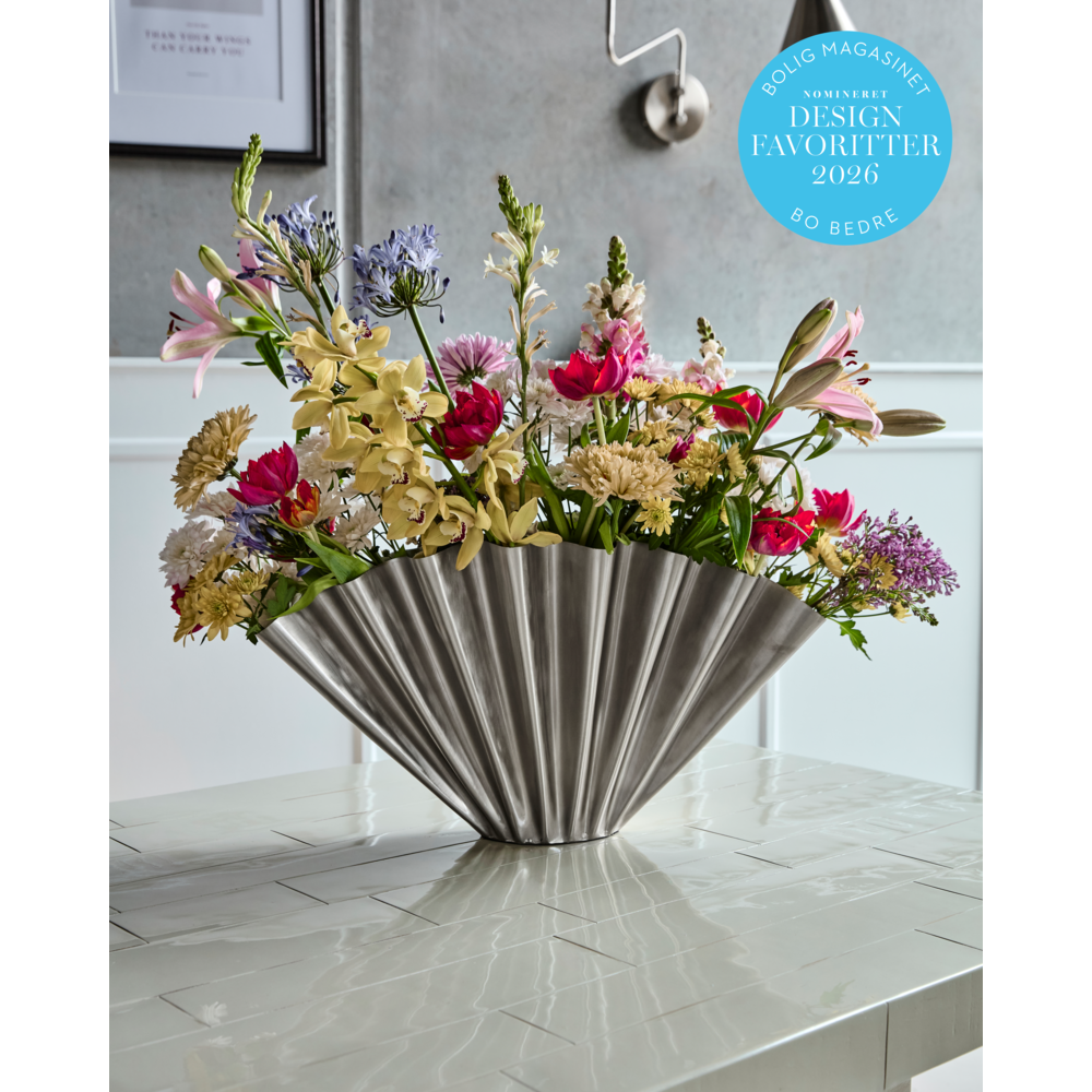 The House Doctor Vase, Flood, in a brushed silver finish displays vibrant flowers on a tiled table in a bright, stylish room. A blue sticker in the corner notes its design award nomination.