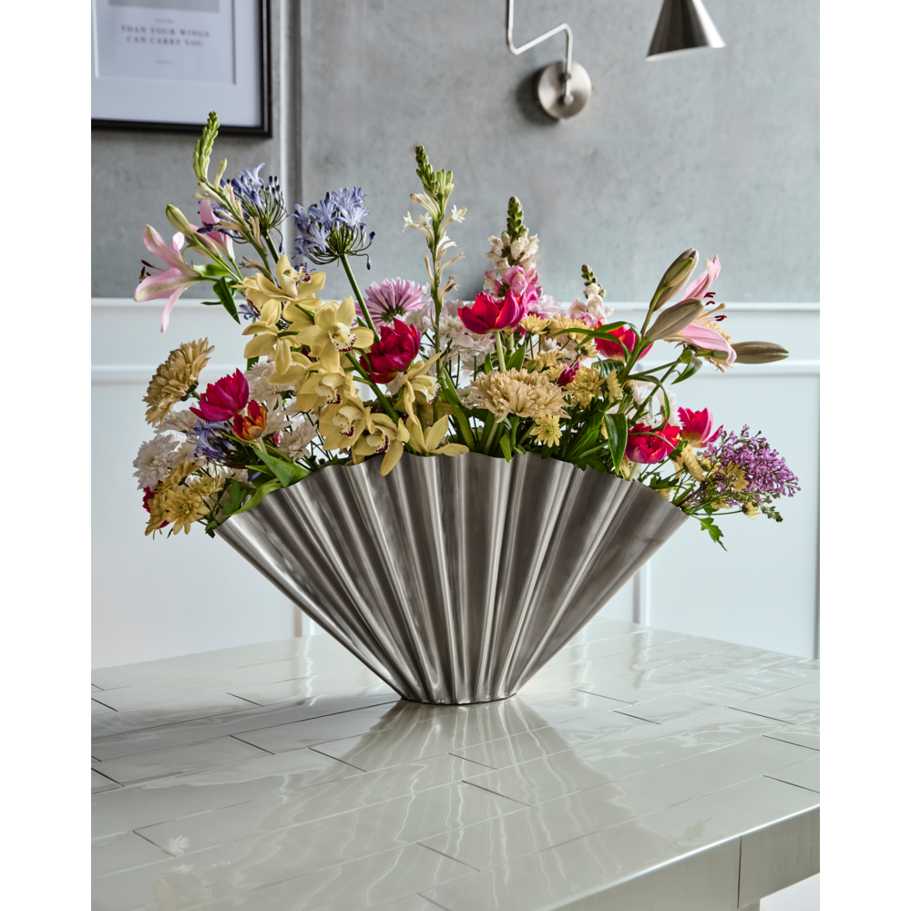 The House Doctor Vase, Flood, in a brushed silver finish holds vibrant pink, yellow, and purple flowers on a light table, bringing modern sculptural style to a contemporary room.