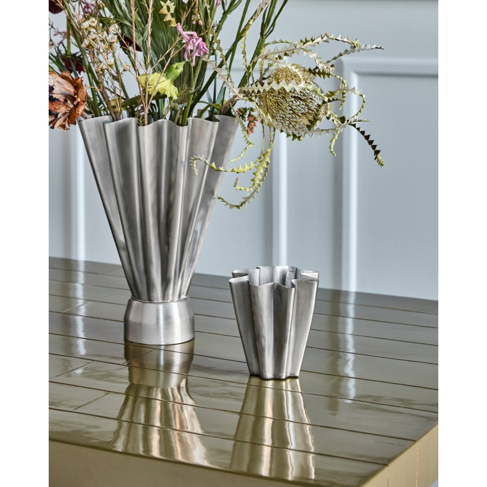 Two House Doctor Vase, Flood, Brushed Silver Finish, Small vases in a fan shape rest on a shiny olive-green table; the taller holds assorted flowers while the other is empty. A light blue paneled wall provides a stylish modern backdrop.