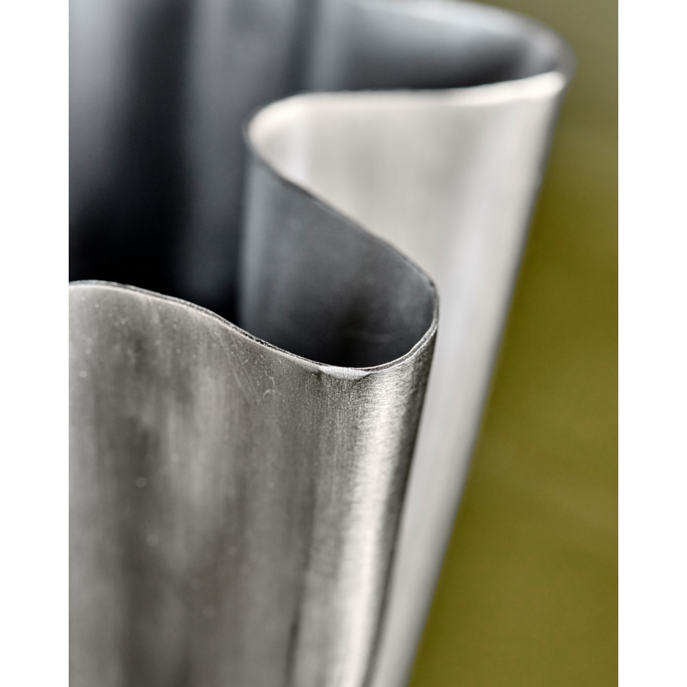Close-up of the House Doctor Vase, Flood, Small with a brushed silver finish and smooth curves, its slightly reflective surface set against a blurred green background—an ideal piece for modern home décor.