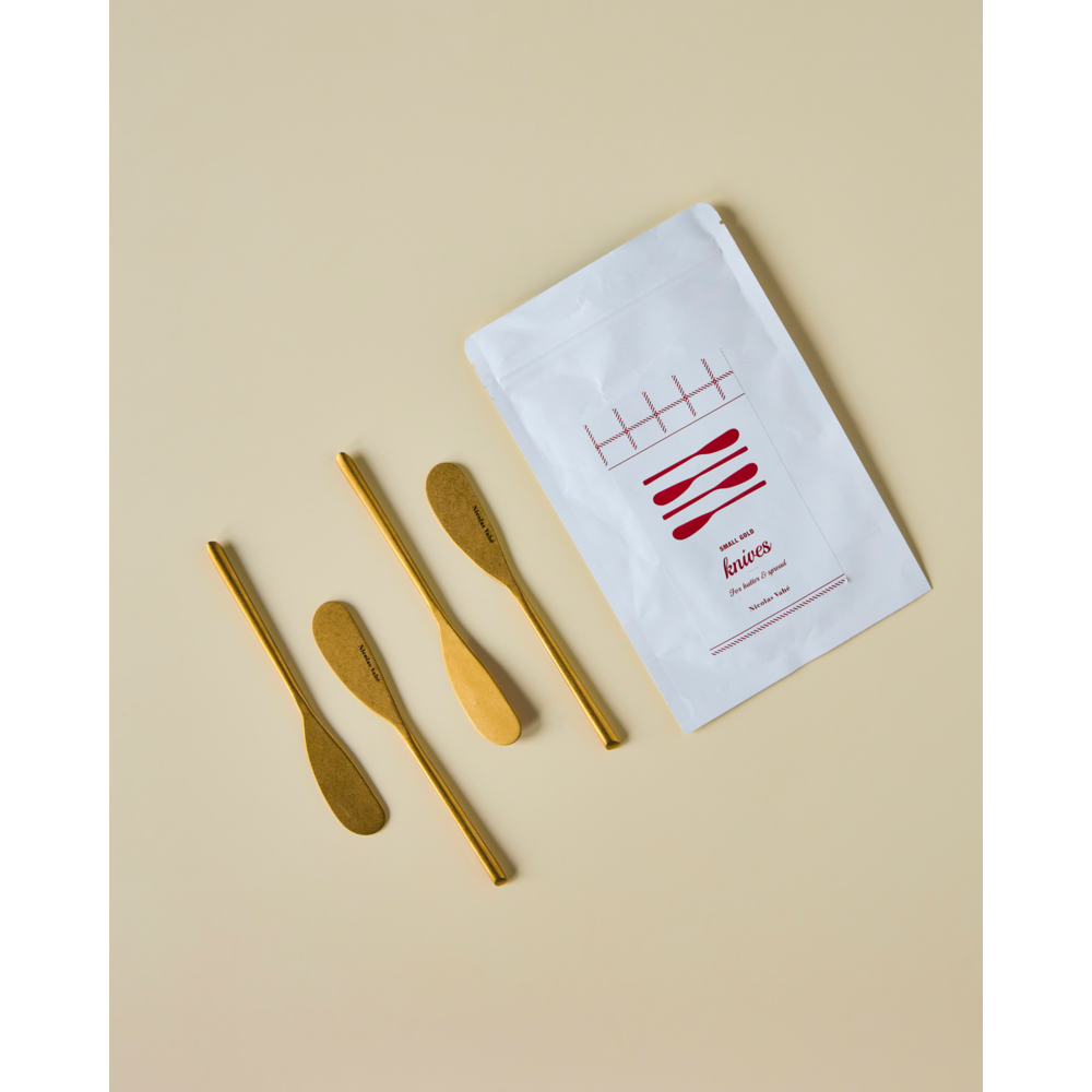 A set of four Nicolas Vahé butter knives in an antique gold finish is displayed beside a white paper packet labeled "knives" with a red graphic, all on a light beige background.