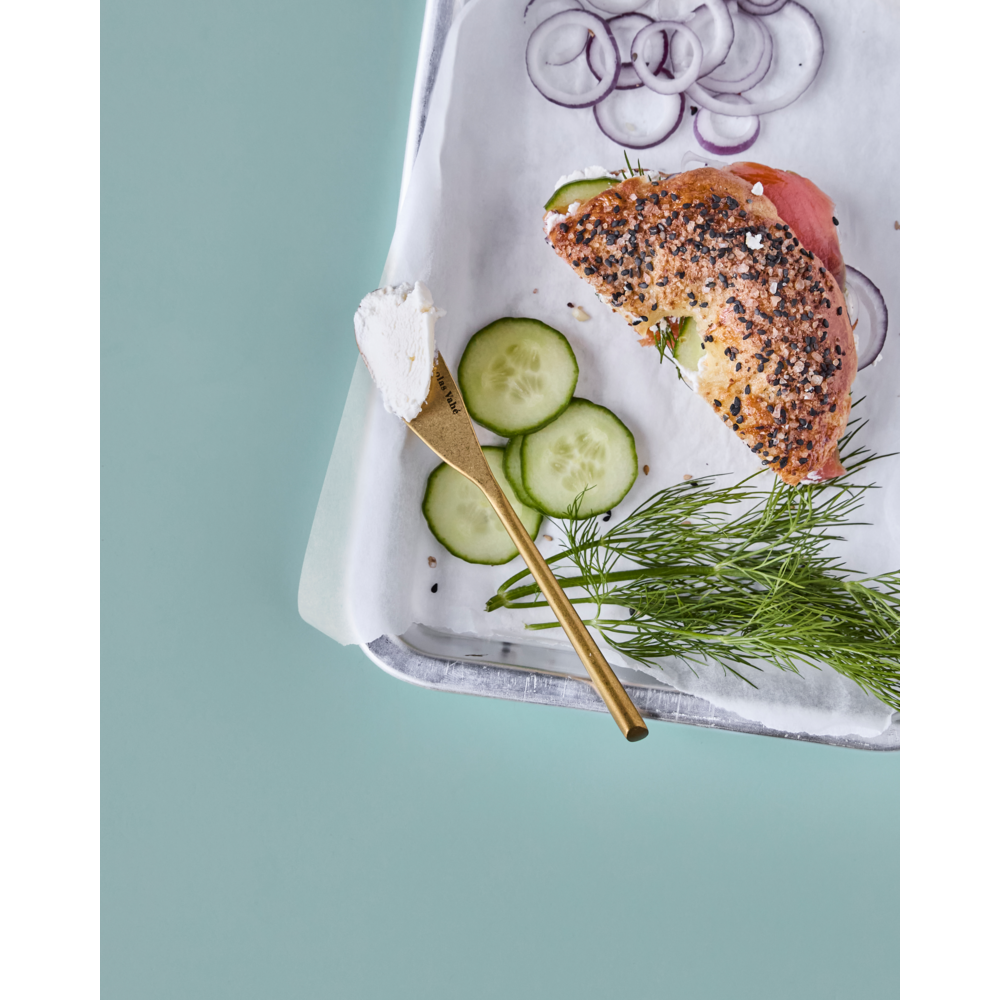 A seeded bagel sandwich with cucumber and meat sits on parchment next to cucumber, dill, red onion, and cream cheese. An antique gold butter knife from the Nicolas Vahé Butter Knives Set (antique gold finish, set of 4) adds elegance against a light blue background.