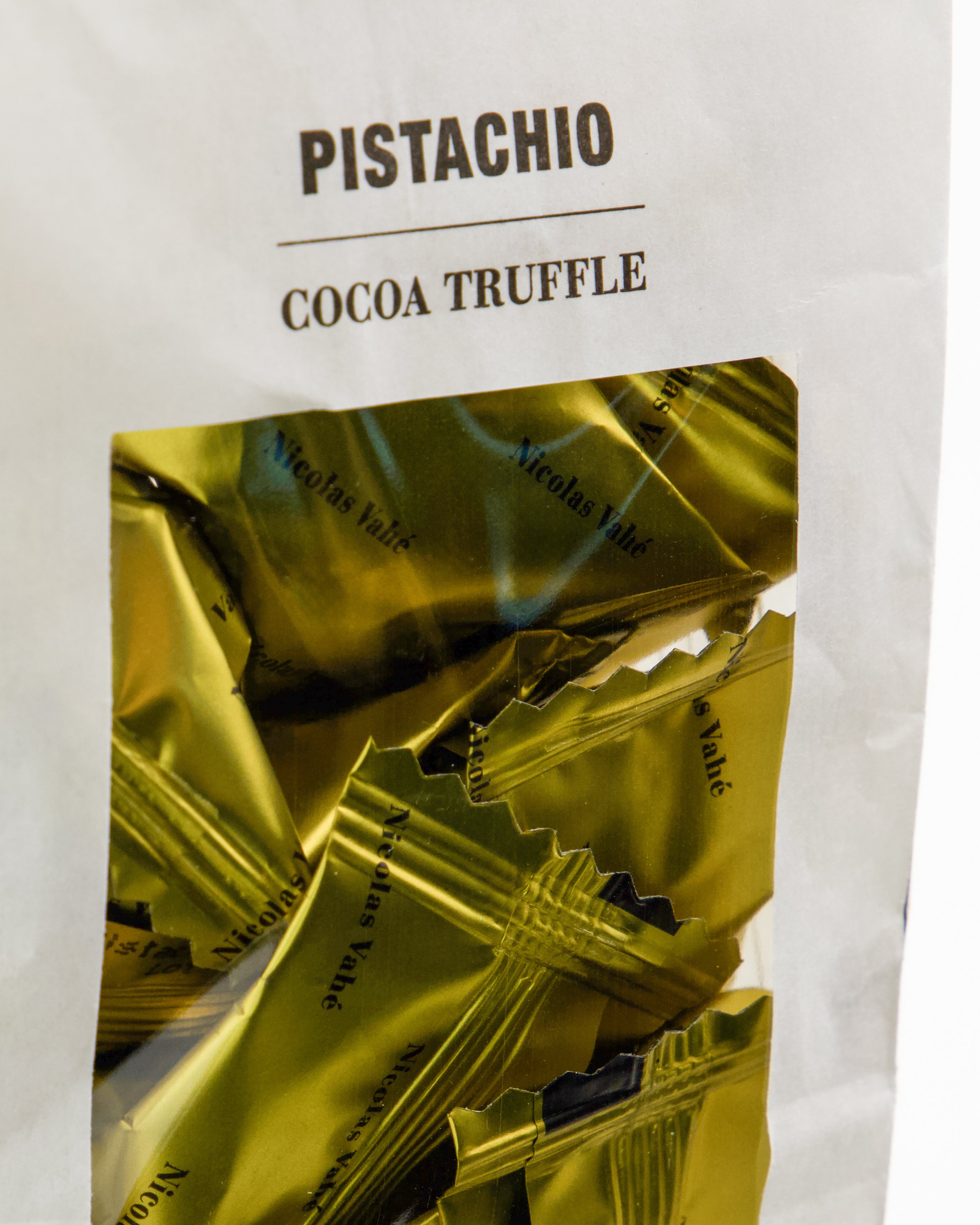 A close-up of a white bag labeled "Cocoa Truffle, Pistachio" by Nicolas Vahé, featuring a clear window that reveals individually wrapped gold candies inside—an ideal treat for anyone who loves the sweet harmony of pistachio and cocoa truffles.