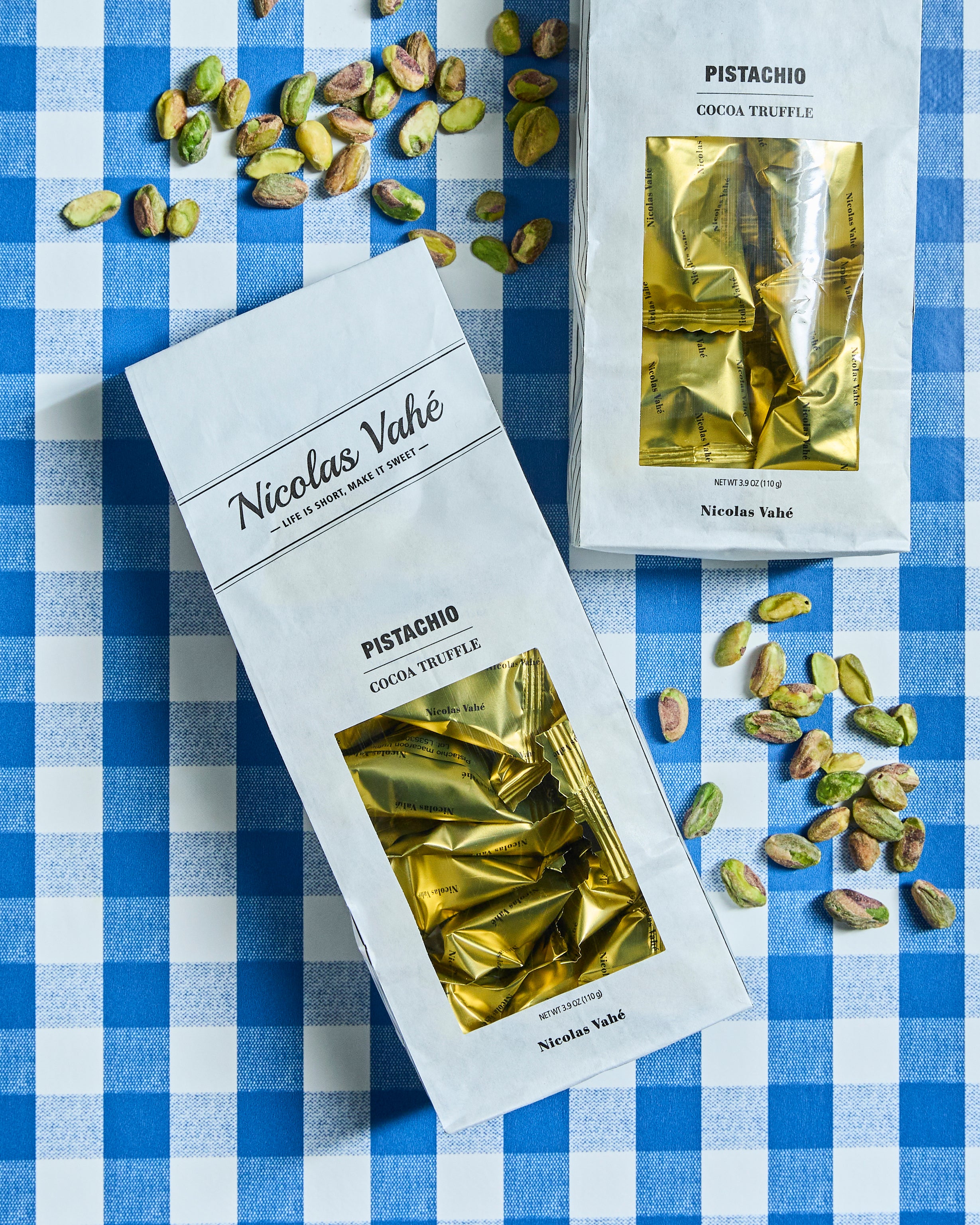 Two boxes of Nicolas Vahé Cocoa Truffle, Pistachio sit atop a blue checkered tablecloth, surrounded by scattered pistachios. Each box’s clear window displays the gold-wrapped truffles inside, creating an inviting scene that pairs beautifully with almond meringue desserts.