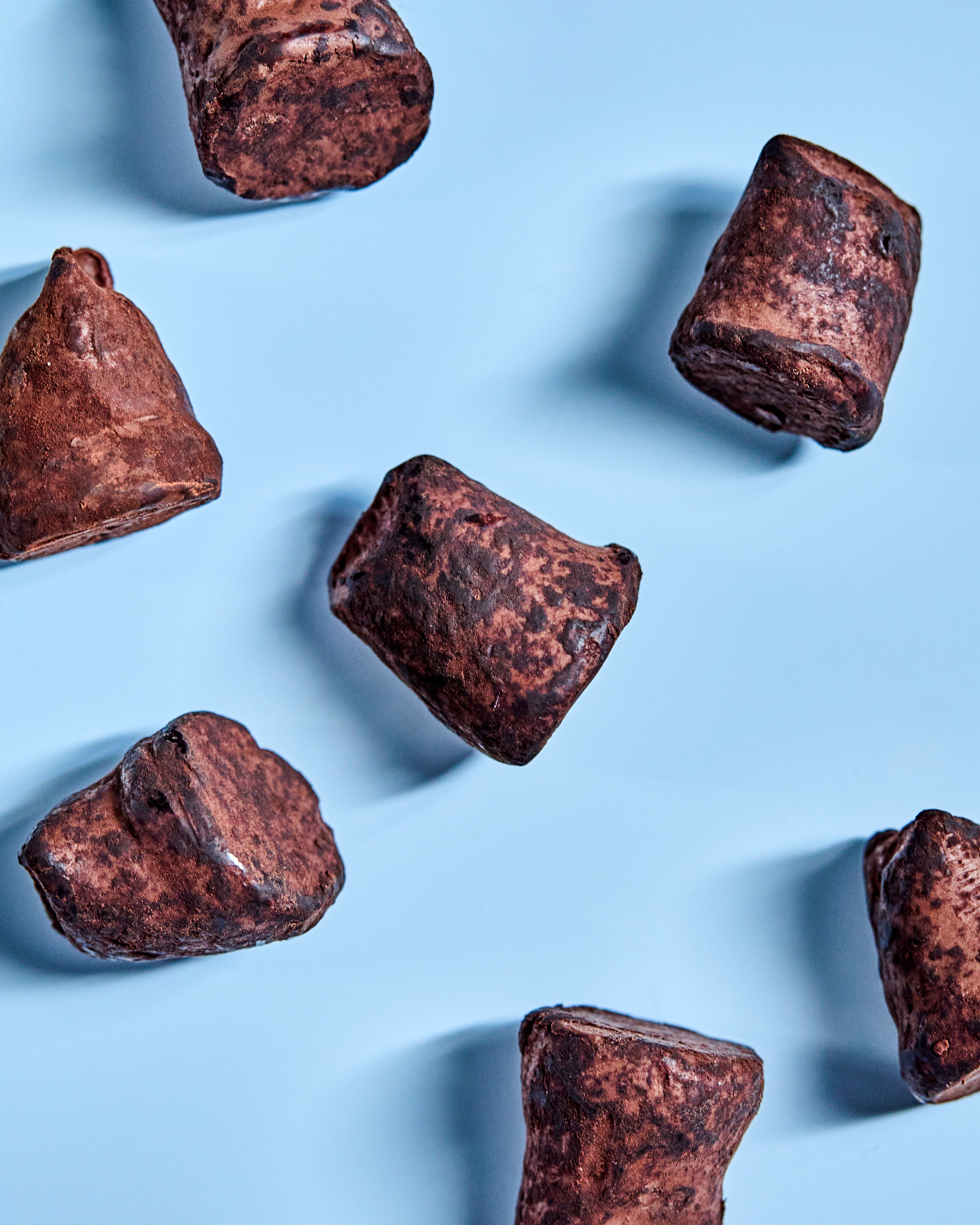 Seven irregularly shaped Nicolas Vahé Cocoa Truffle, Pistachio confections are scattered on a light blue background, each dusted with cocoa powder and casting soft shadows, evoking the delicate texture of almond meringue and the vibrant hue typical of pistachio treats.