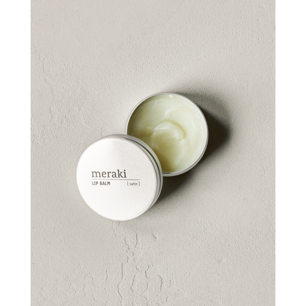 An open round white tin of Meraki Organic Lip Balm, Satin rests on a light textured surface. The tilted lid displays “meraki LIP BALM [ satin ].” The pale balm inside is enriched with natural oils.