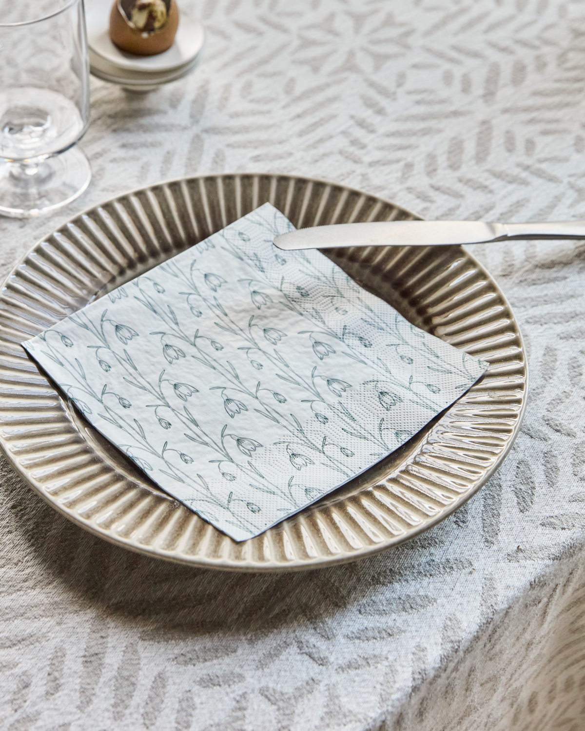 The House Doctor Snowdrop Napkins in White & Green (Pack of 40) are displayed on a decorative plate with a knife, set atop a textured tablecloth—perfect for seasonal table settings, alongside a glass and a small chocolate dish.