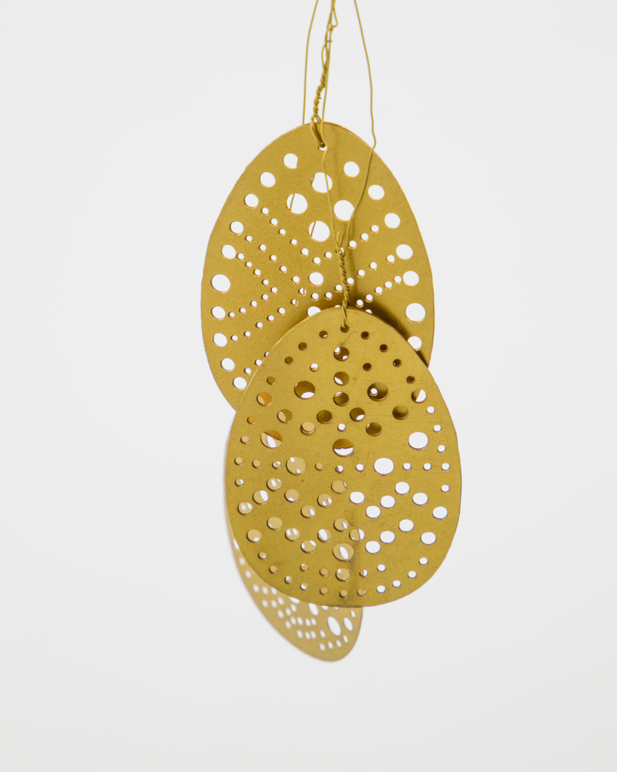 The Set of 3 Easter Eggs in Brass, Cut by House Doctor features overlapping, egg-shaped ornaments with circular cut-out patterns, suspended by thin gold threads against a plain white background.