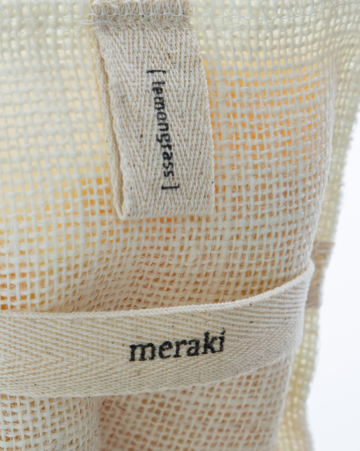 A close-up of a beige jute bath mitt filled with lemongrass from Meraki, visible through mesh fabric packaging with two labels: "Bath Mitt, in Jute Filled with Lemongrass" and "Meraki.