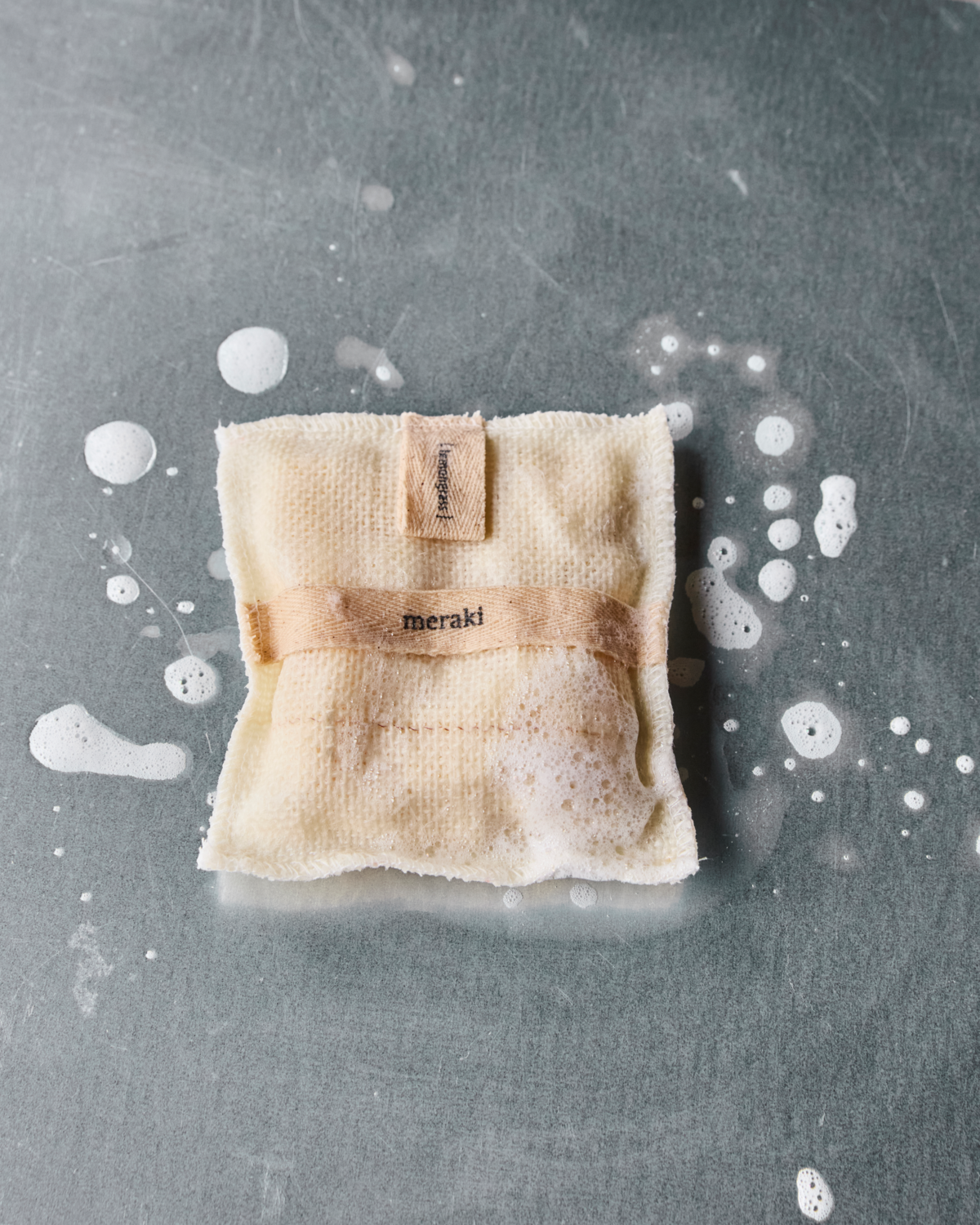 A small, beige jute Bath Mitt filled with lemongrass from Meraki rests on a gray surface among bubbles—ideal for exfoliating and revitalizing your skin during your bath routine.
