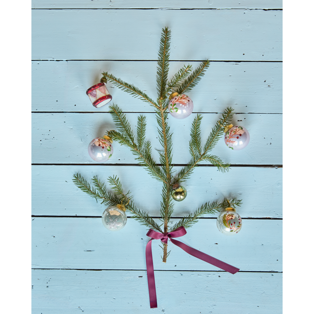 A pine branch tied with a purple ribbon and decorated with six Christmas Ornament Baubles Set of 4, Antique Pink Handpainted, including a pink glass ornament, arranged on a light blue wooden surface.