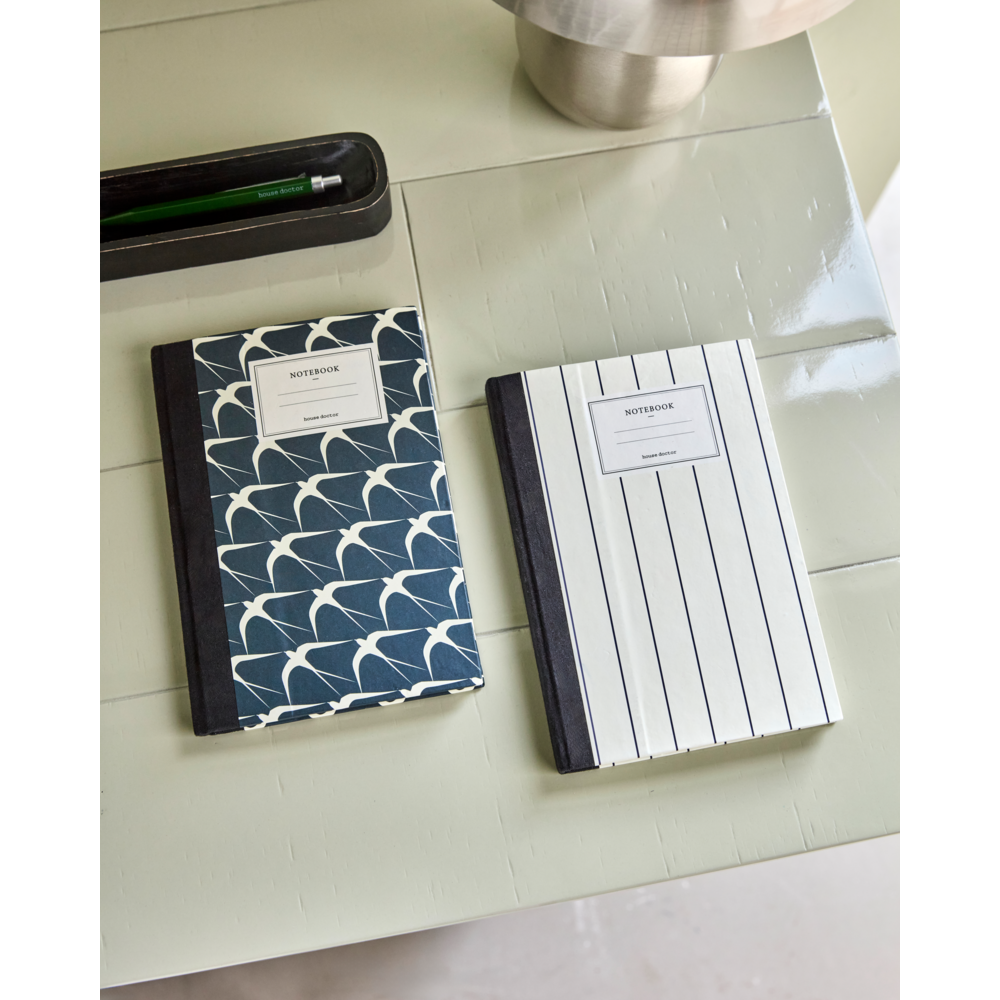 The House Doctor "Notebook / Journal, Sand & Birds" set of 2 features stylish covers—one blue with a white wave pattern and one with black vertical stripes—resting on a light green table, paired with a pen and holder for daily planning.