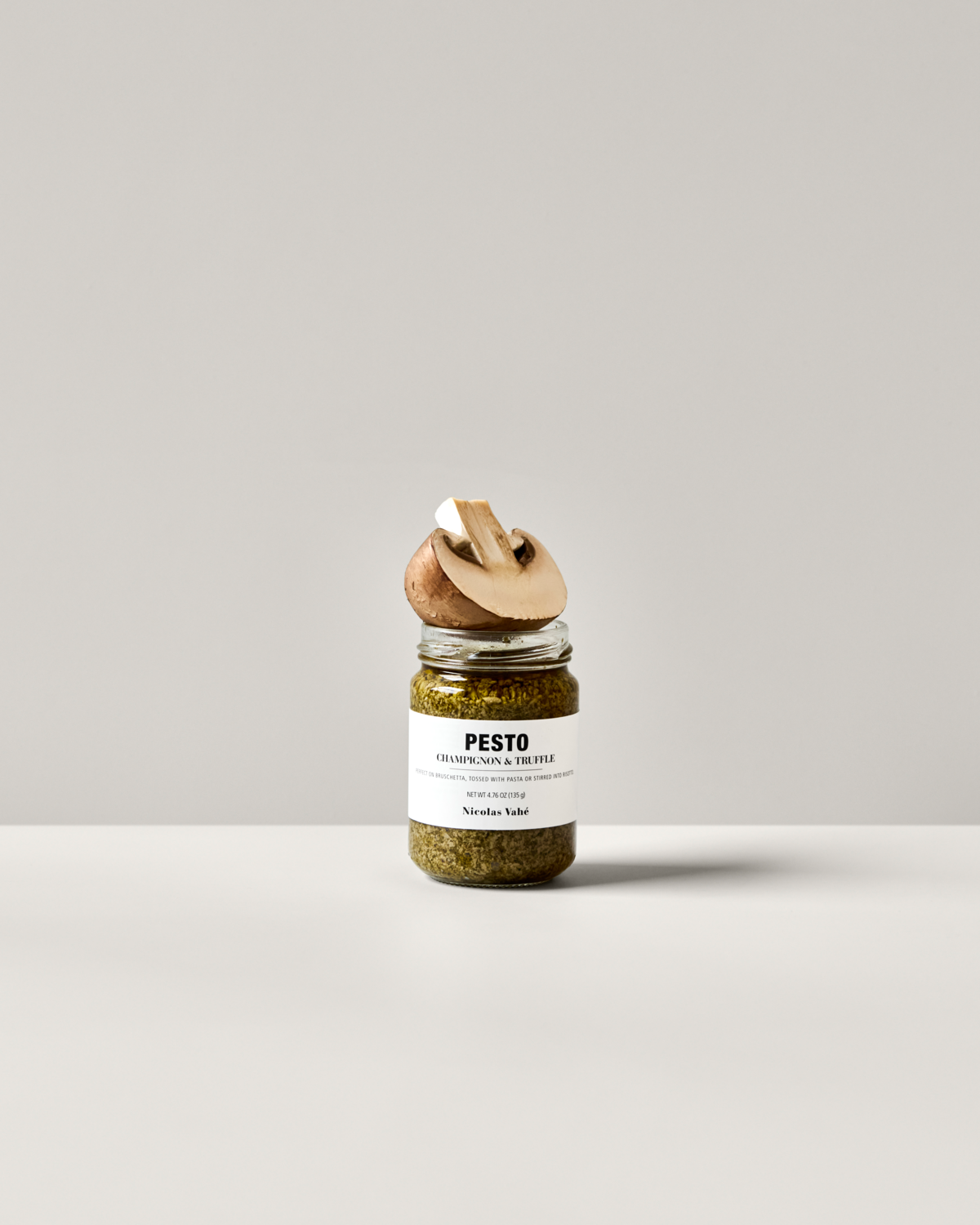 A jar of Nicolas Vahé Pesto, Champignon & Truffle with a white label sits on a white surface against a light gray background. A whole mushroom and a slice are balanced on the lid.