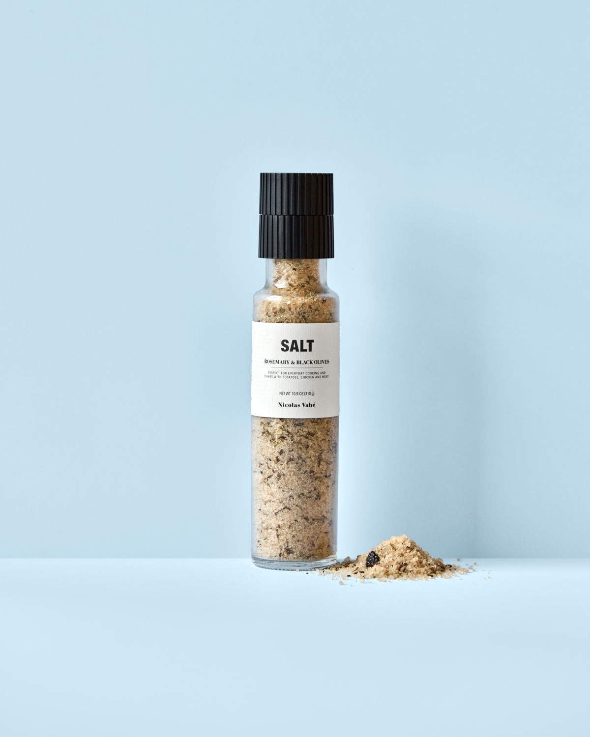 A clear glass grinder of Nicolas Vahé Salt, Rosemary & Black Olives stands upright on a light blue surface, with a small pile of the aromatic salt blend spilled to its right. The grinder features a black top and a white label with text.