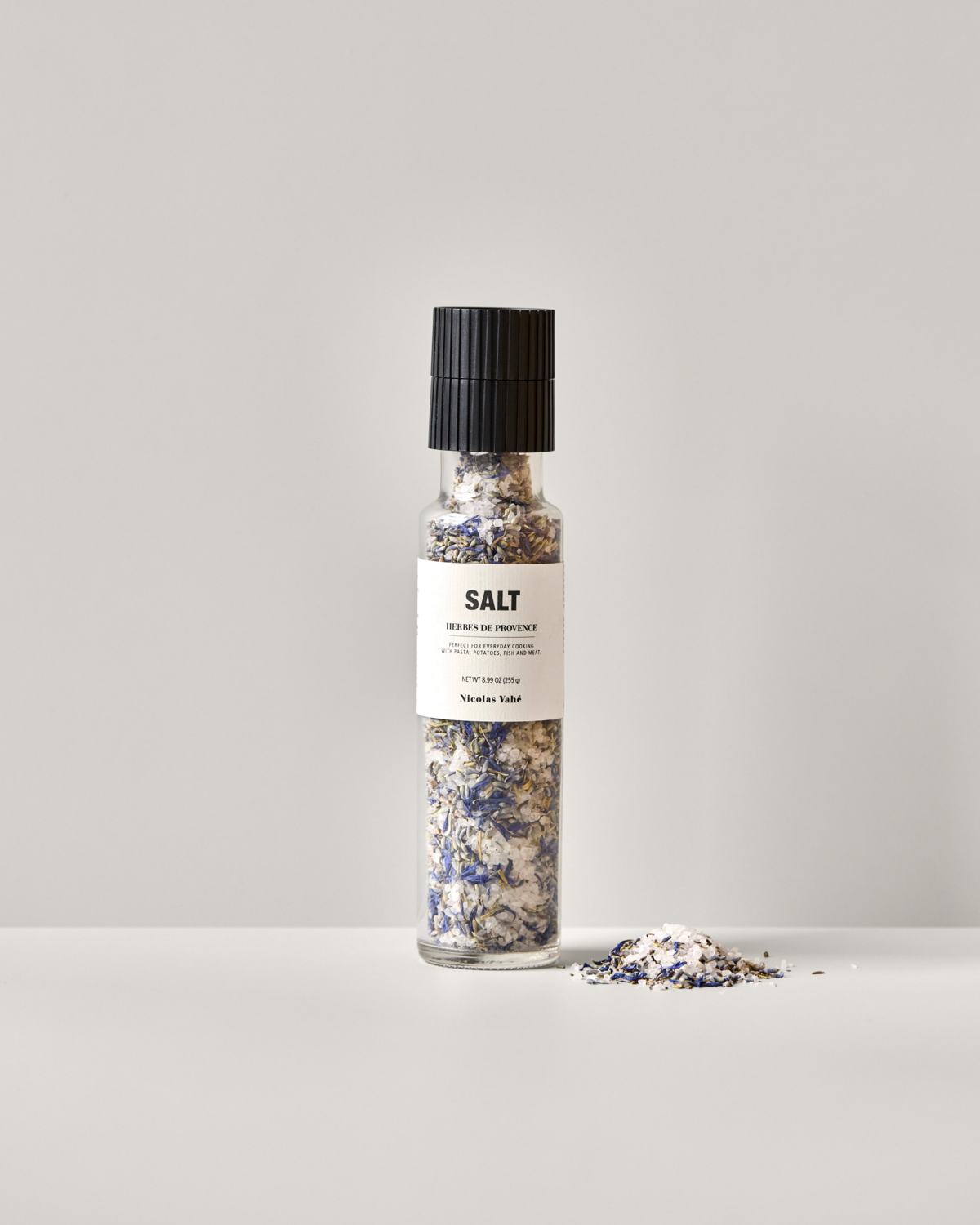 A clear glass grinder of Nicolas Vahé Salt, Herbes de Provence rests on a light surface, with some sea salt and herb mix containing rosemary scattered nearby. The label reads SALT HERBS DE PROVENCE.