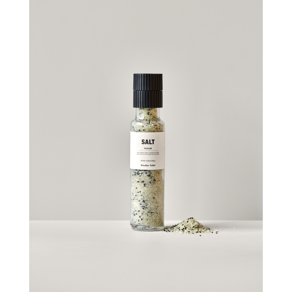 A clear Nicolas Vahé Salt, Wasabi grinder, filled with white salt and black flecks, sits on a white surface. Next to it is a small pile of wasabi salt mixed with black sesame seeds. The grinder’s label reads "Salt, Wasabi.