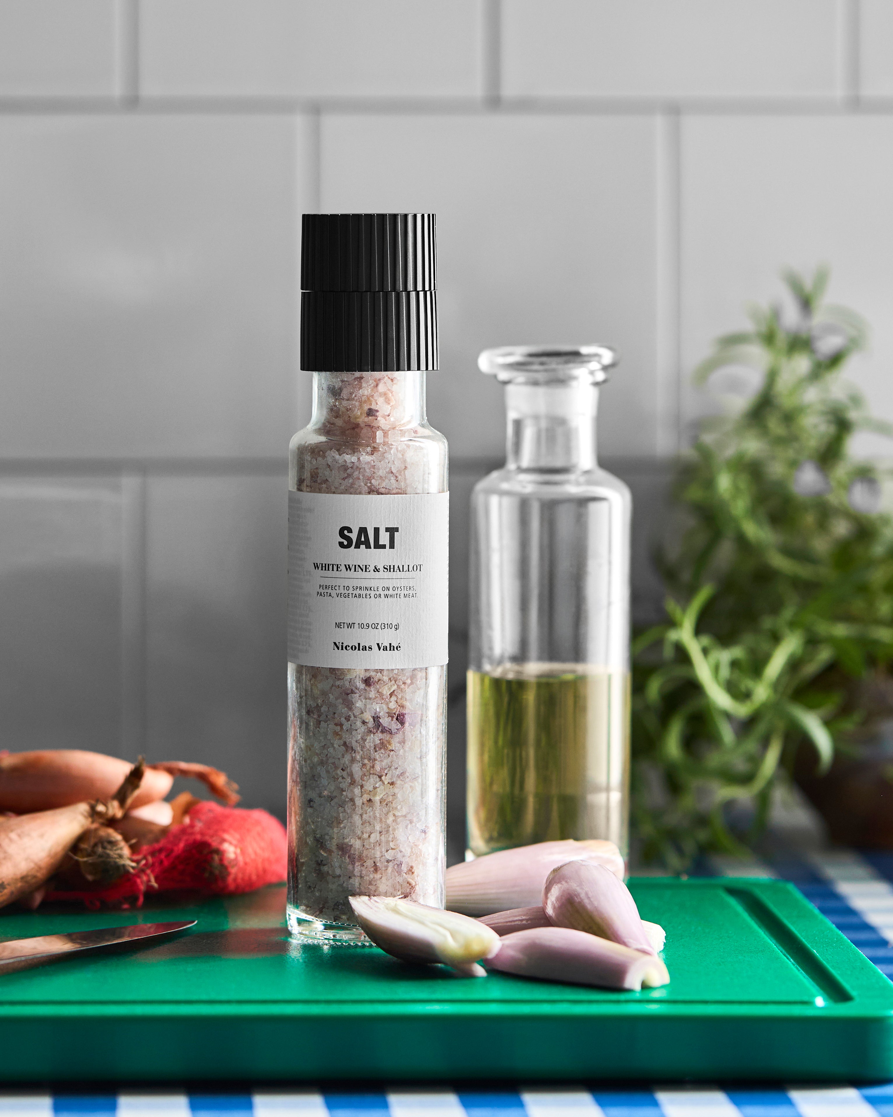 A Nicolas Vahé Salt, White Wine & Shallot grinder rests on a green cutting board along with fresh shallots, a knife, a jar of liquid, and aromatic herbs. Set against white tiled kitchen walls, the scene highlights the unique blend of sea salt, fragrant shallots, and subtle white wine notes in this gourmet seasoning.