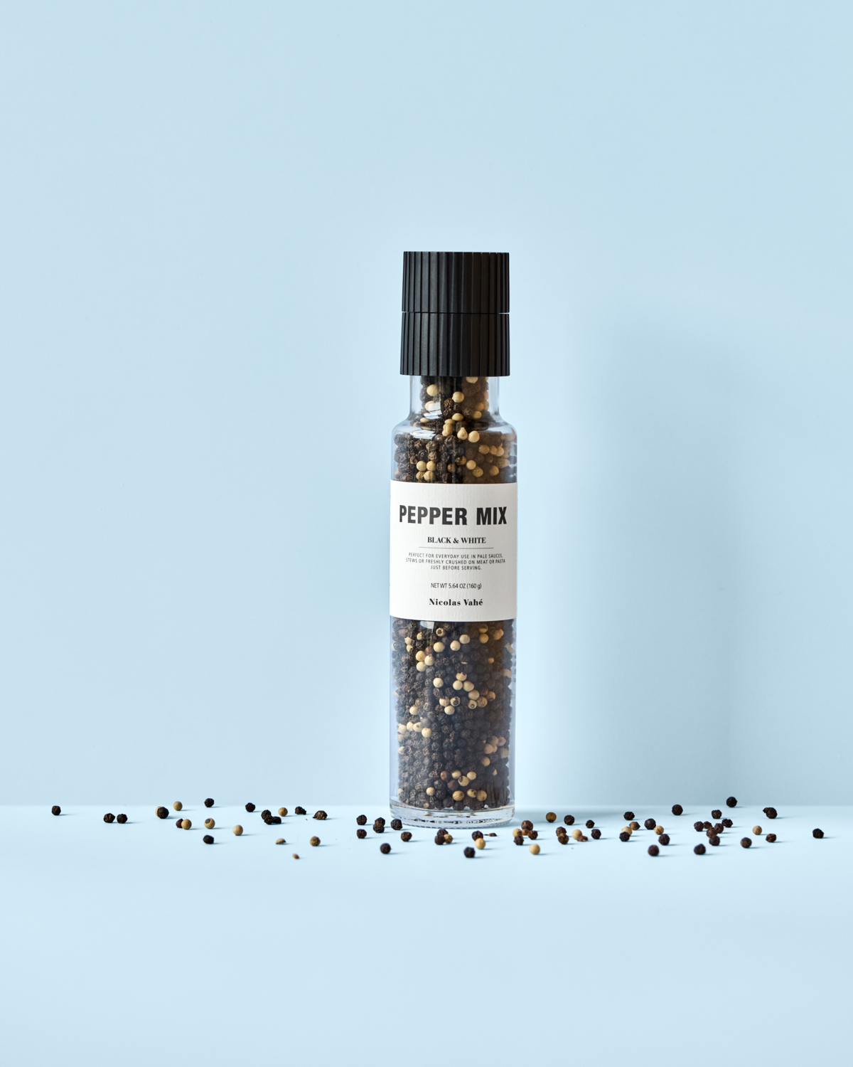 A Nicolas Vahé Pepper Mix, Black & White grinder stands upright on a light blue background, filled with black and white peppercorns plus pink peppercorns, with scattered seasoning around its base.
