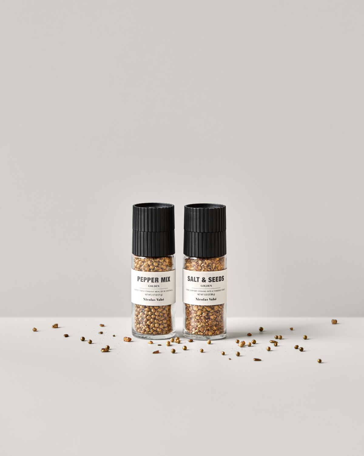 The Nicolas Vahé Gift Box features Golden Pepper Mix and Golden Salt & Seeds grinders, displayed side by side on a white surface with scattered seeds and peppercorns—perfect for adding to any gift box.