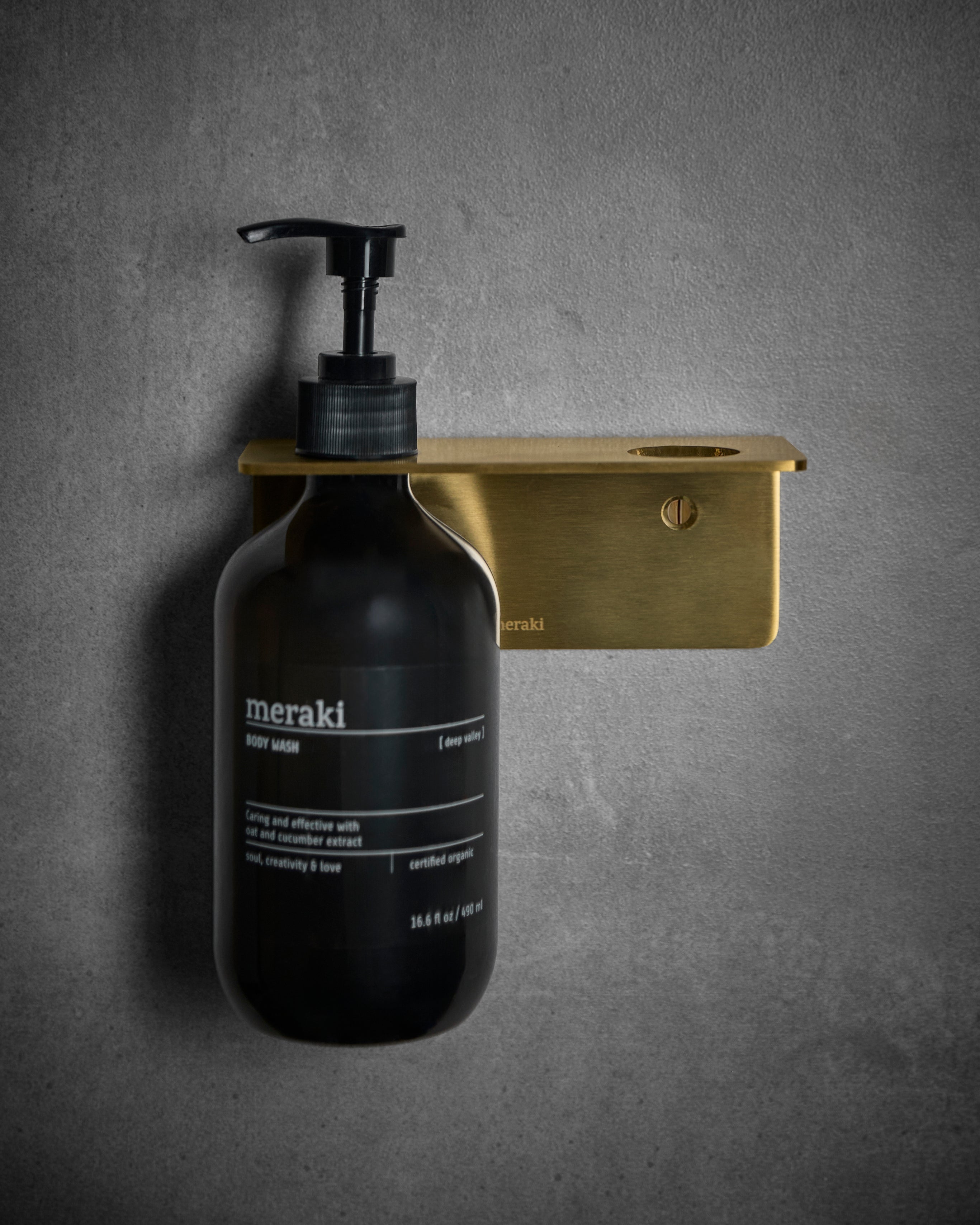 A black Meraki body wash bottle with a pump is displayed on the Meraki Bottle Hanger 2 Holes in a stylish brushed brass finish, mounted against a gray textured wall to provide both elegant decor and practical bathroom organisation.