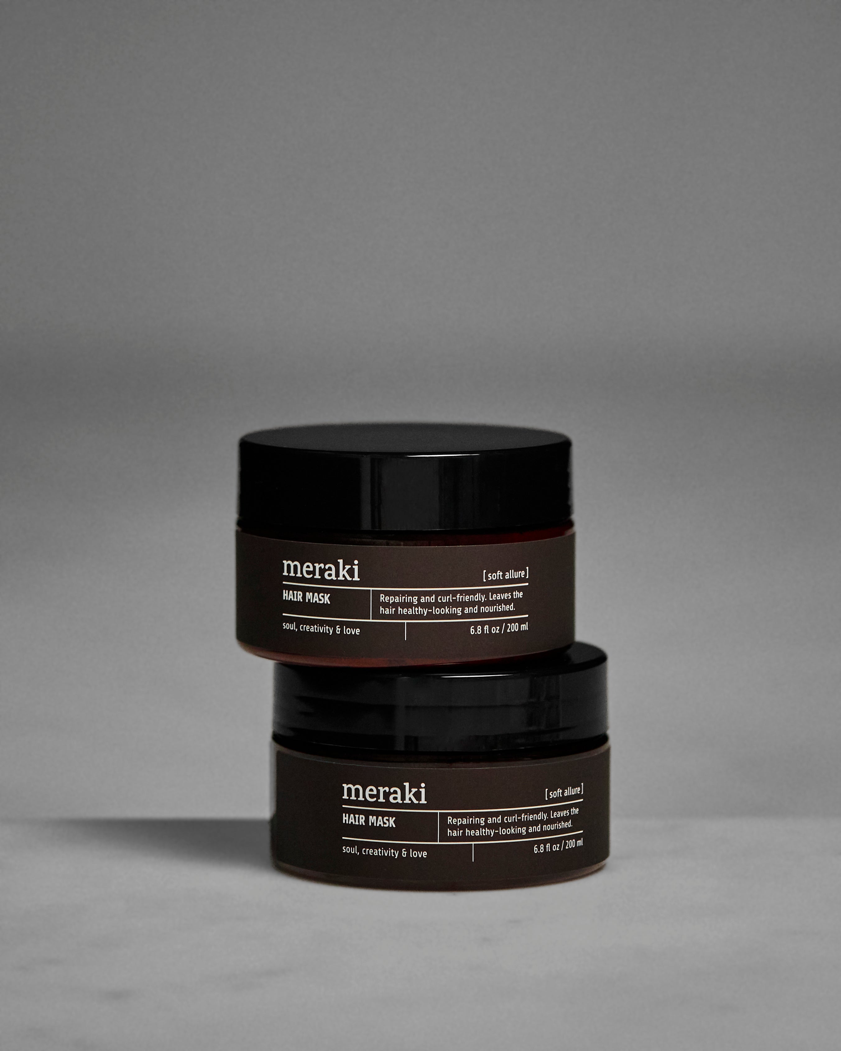 Two black jars of Meraki Hair Mask, Soft Allure, one stacked atop the other, are displayed against a plain gray background. The labels highlight nourishing and restorative benefits for curl-friendly hair care, helping you achieve nourished, healthy hair.