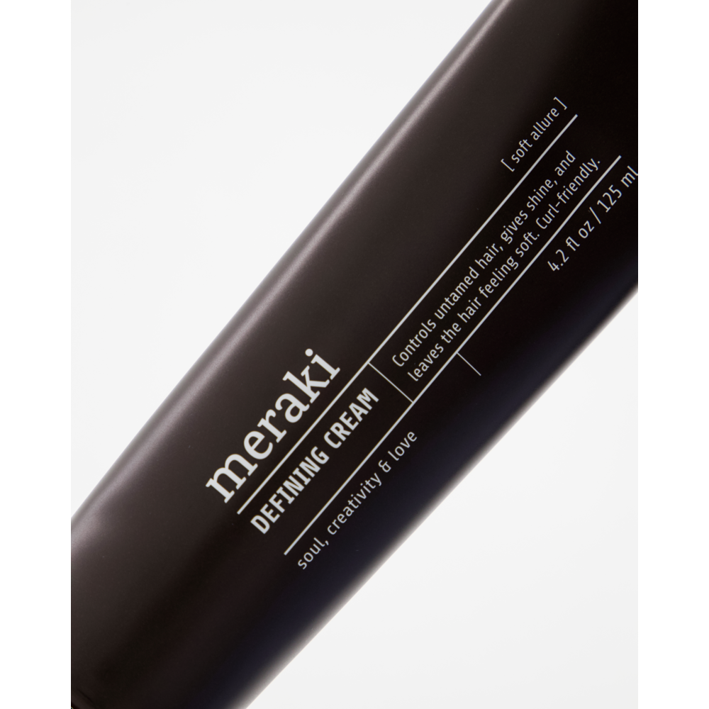 A close-up of a black tube labeled Meraki Defining Cream, Soft Allure. The text describes the product as a nourishing hair cream that tames unruly hair, adds shine, and leaves hair feeling soft. Ideal for curl-friendly hair care routines, the tube contains 4.2 fl oz (125 ml).