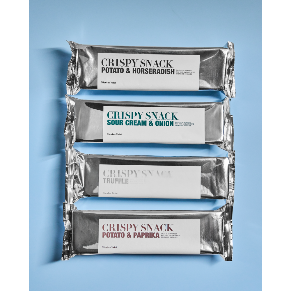 Four sleek silver packages of Nicolas Vahé Crispy Snack—featuring Potato & Horseradish, Sour Cream & Onion, Truffle, and Potato & Paprika flavors—are stacked vertically against a light blue backdrop. Experience the perfectly crunchy texture and bold taste of these gourmet potato snacks, including the indulgent Truffle variety.