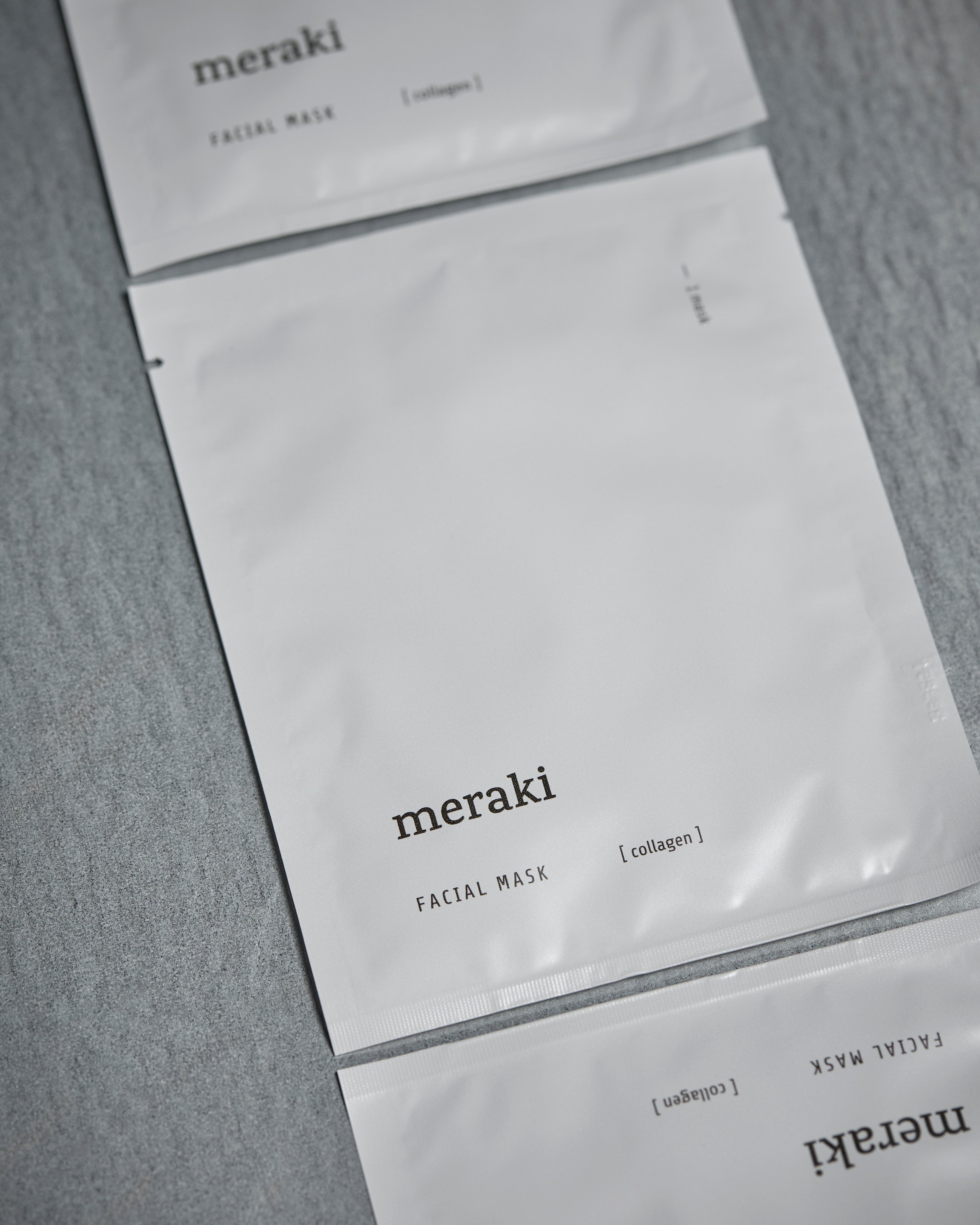 Three flat, white packets of Meraki Face Mask, Collagen—designed to boost skin elasticity with potent marine collagen—are arranged on a gray surface, with two fully visible and one partially visible at the top and bottom of the image.