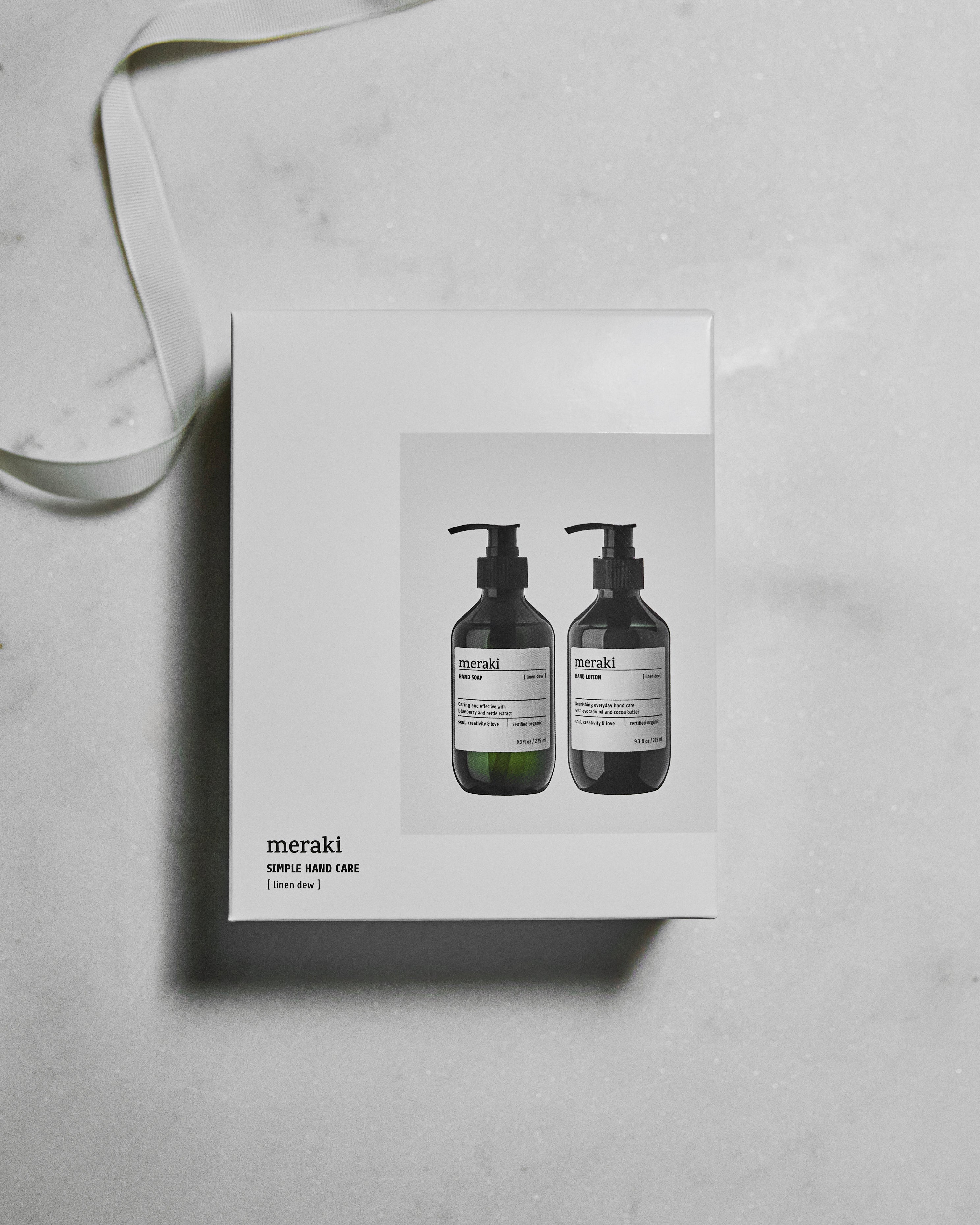 A white Meraki Giftbox Linen Dew Organic—containing hand soap and hand lotion—is displayed with an image of the two pump bottles on the front. The box sits on a light marble surface, with a white ribbon to its left.