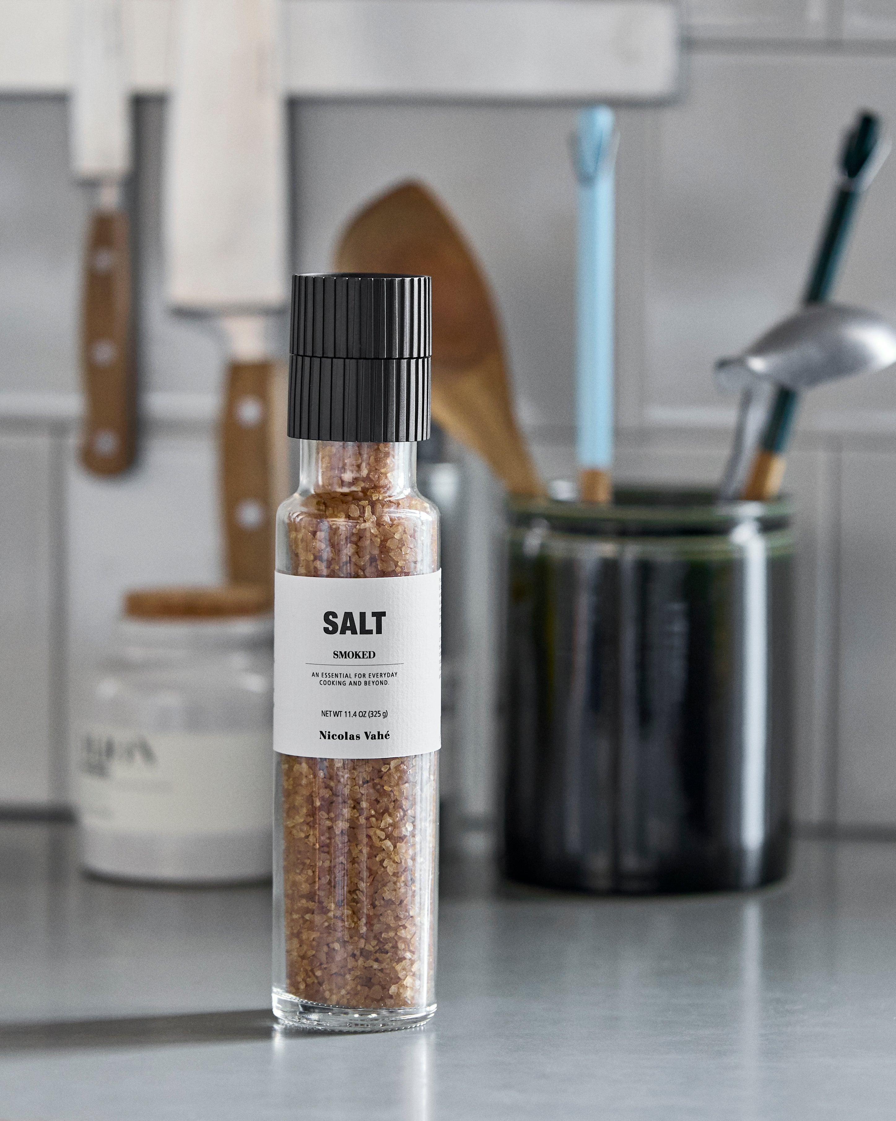 A glass grinder of Nicolas Vahé Smoked Salt rests on a kitchen counter. In the softly blurred background, various kitchen utensils, a knife, a dark jar, and a white container can be seen.