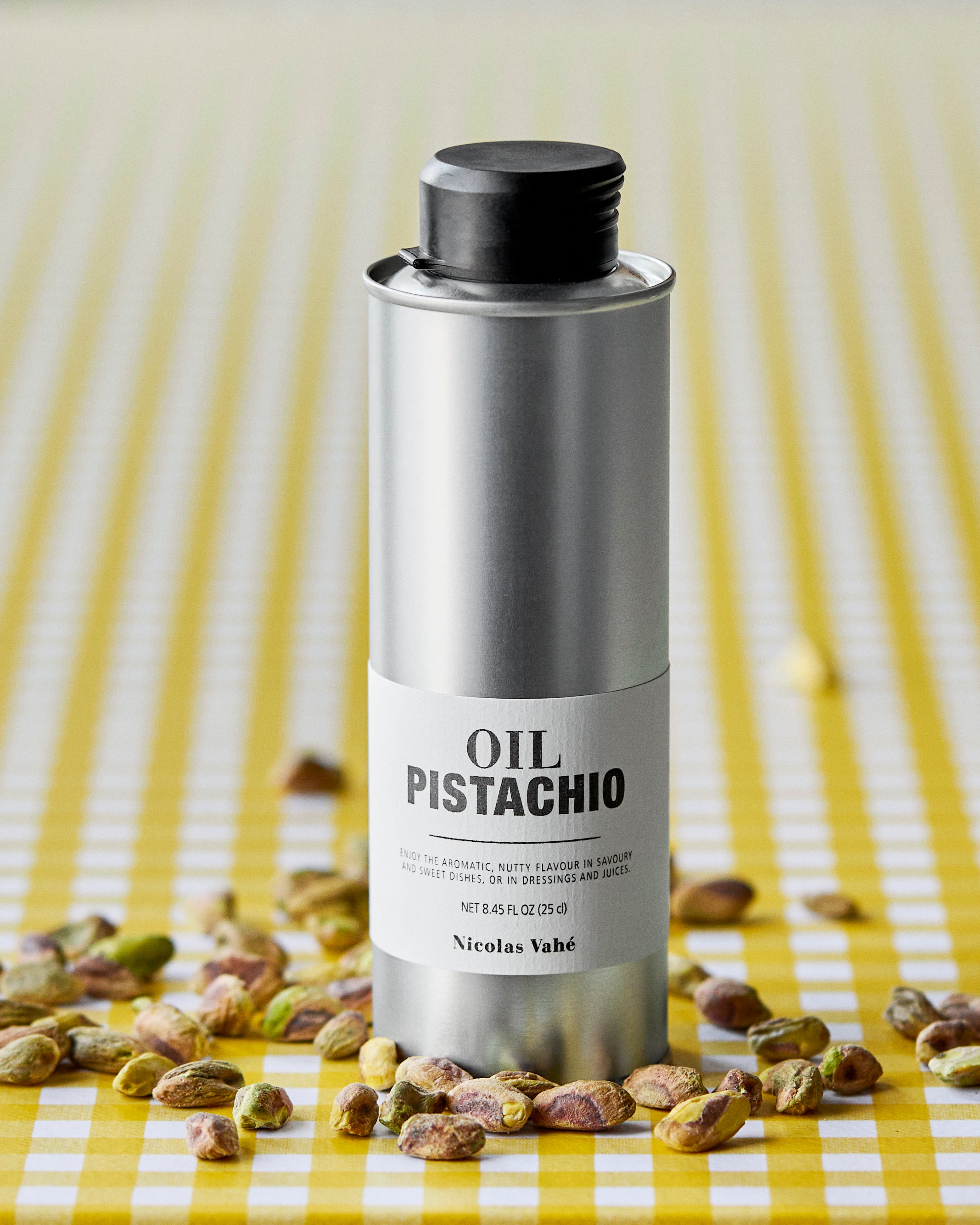 A silver tin of Nicolas Vahé Pistachio Oil rests on a yellow and white checkered surface, surrounded by scattered pistachios, inviting you to elevate your dishes with its rich, nutty flavor.