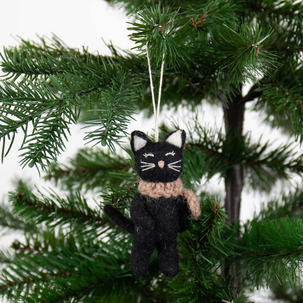 A Black Cat Christmas Ornament, Handmade in Wool, featuring a black cat in a tan scarf hanging from a Christmas tree branch.