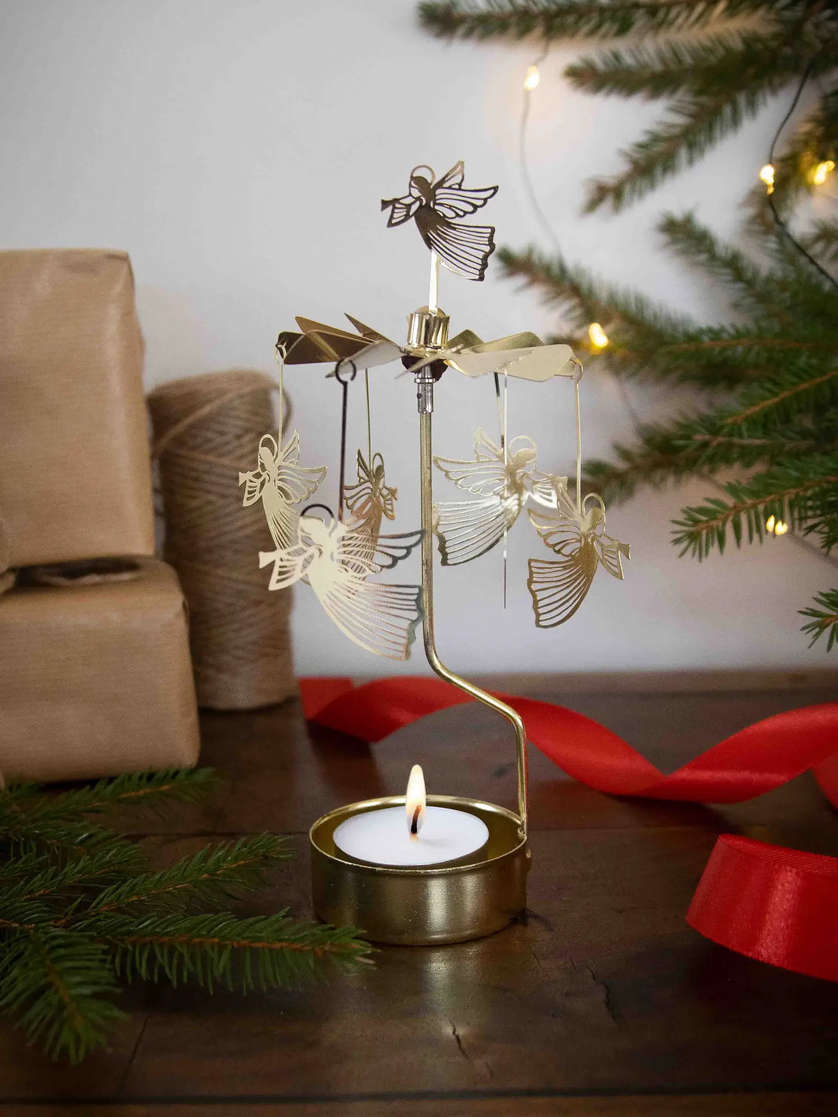 A Tealight Spinner, Flying Angel in Gold by Pluto Design Sweden spins gracefully atop a lit tealight, surrounded by wrapped gifts, twine, evergreen branches, and a red ribbon on a wooden table.
