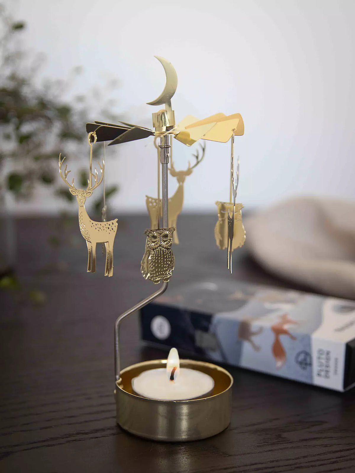 The “Tealight Spinner, Winter Night in Gold” by Pluto Design Sweden is an enchanting home decor accent, featuring delicate hanging deer, owls, and a crescent moon that gracefully spin from the warmth of a lit tealight below. Set on a dark table with a blurred backdrop, this gold carousel infuses any space with charm and coziness.