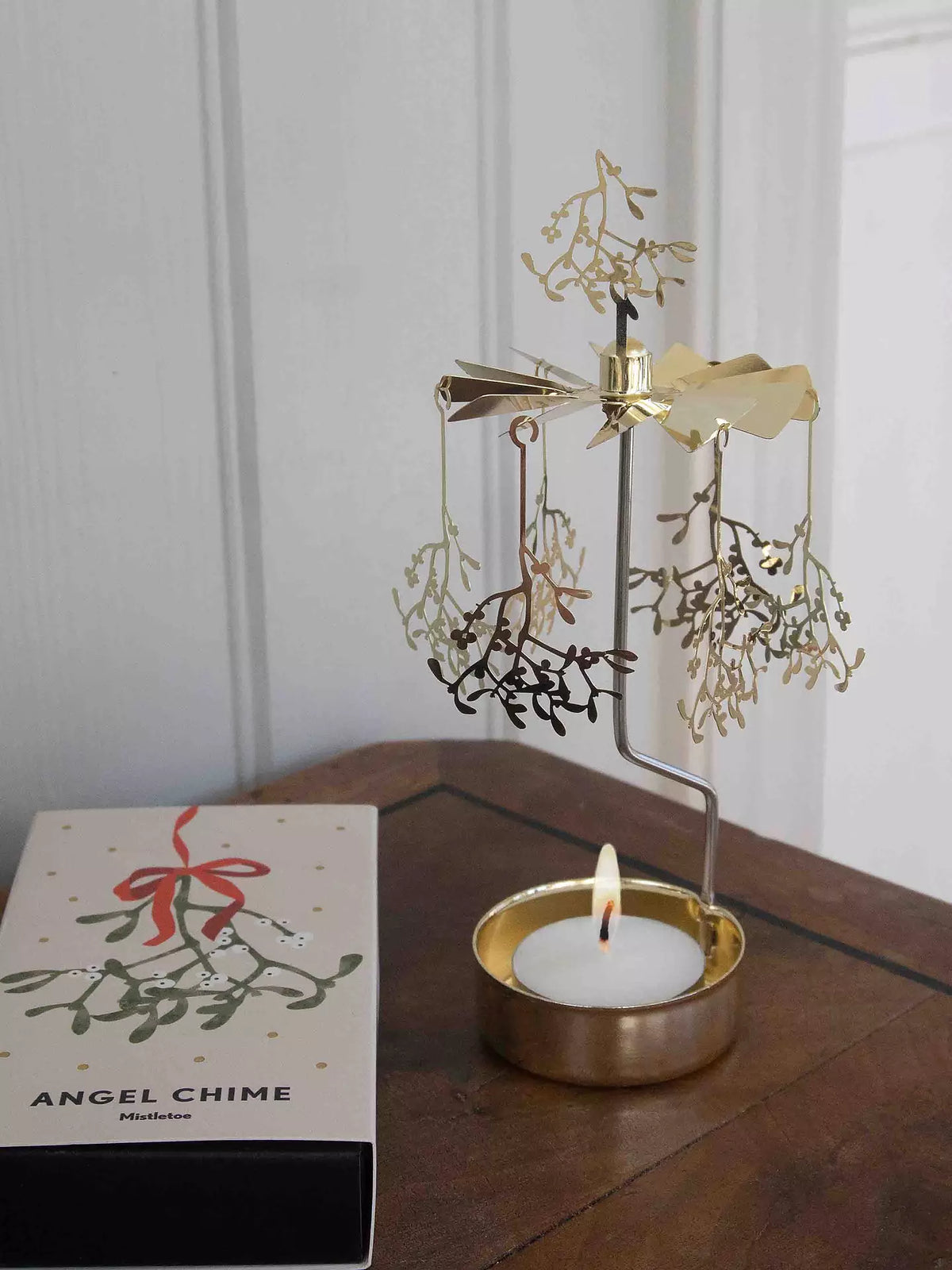 A Tealight Spinner, Mistletoe in Gold from Pluto Design Sweden twirls gracefully above a glowing candle, spreading Christmas charm with its mistletoe-shaped ornaments. Nearby, a box labeled “Angel Chime Mistletoe” rests on a white surface.